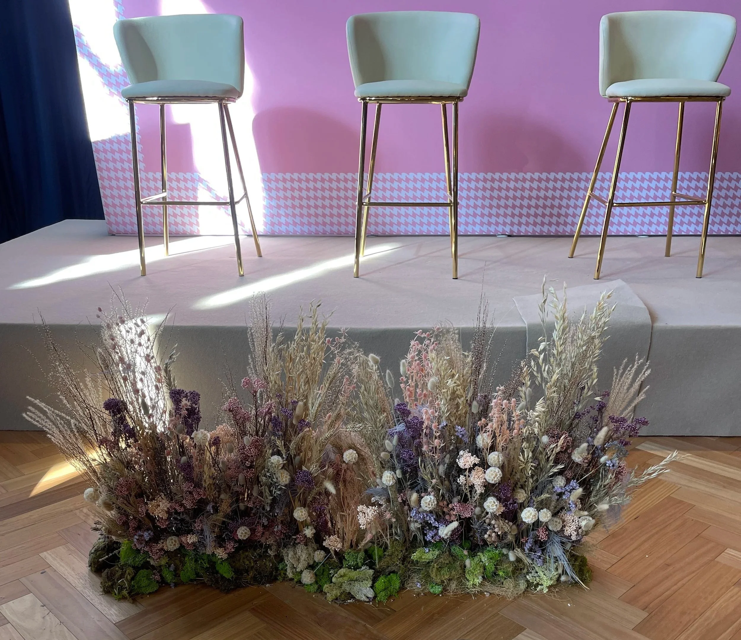 Three pastel green chairs with gold legs on a stage with pink houndstooth backdrop and a floral arrangement in front of the stage.