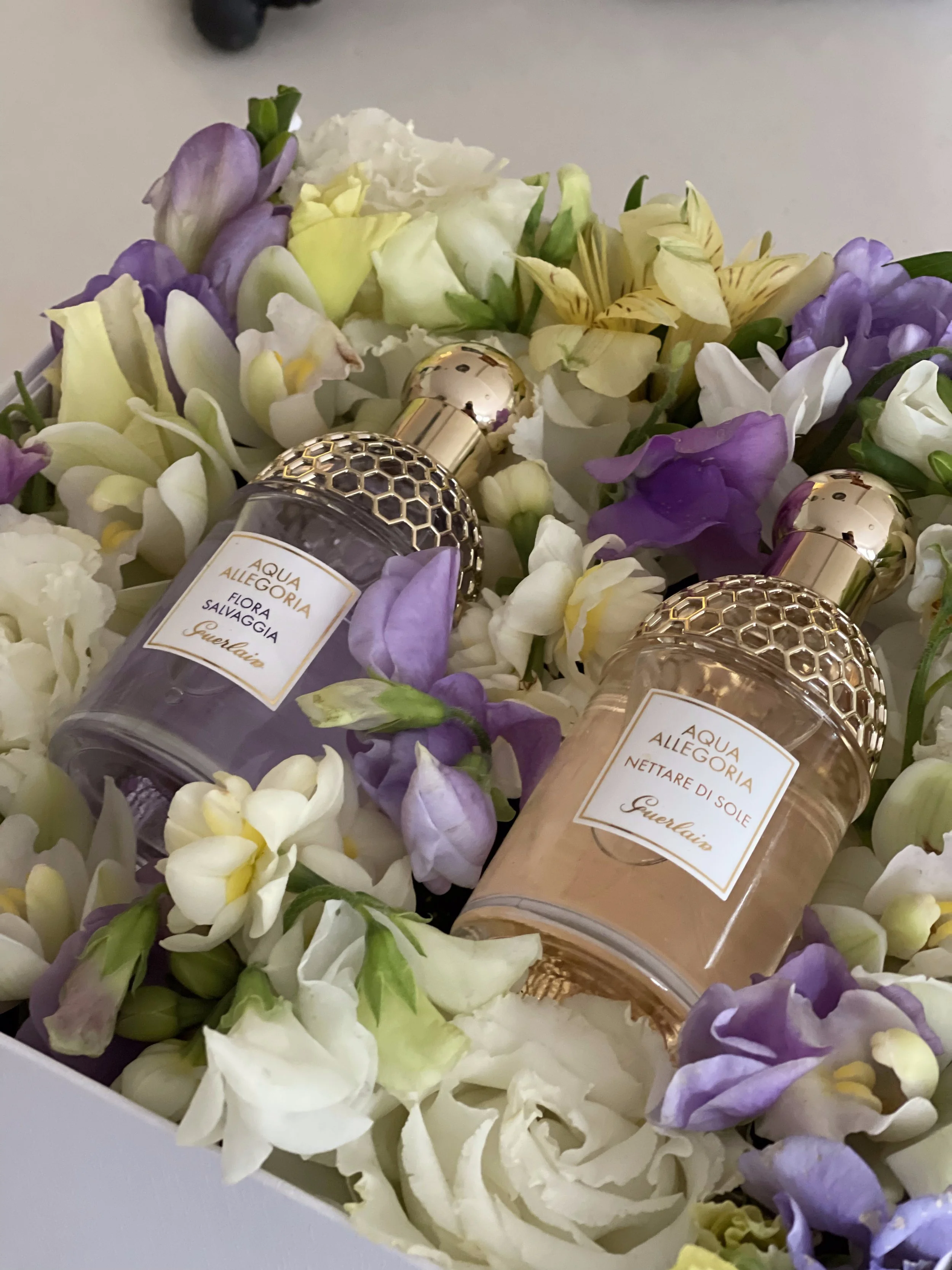 Two bottles of Guerlain Aqua Allegoria fragrances placed among white, purple, and yellow flower petals.