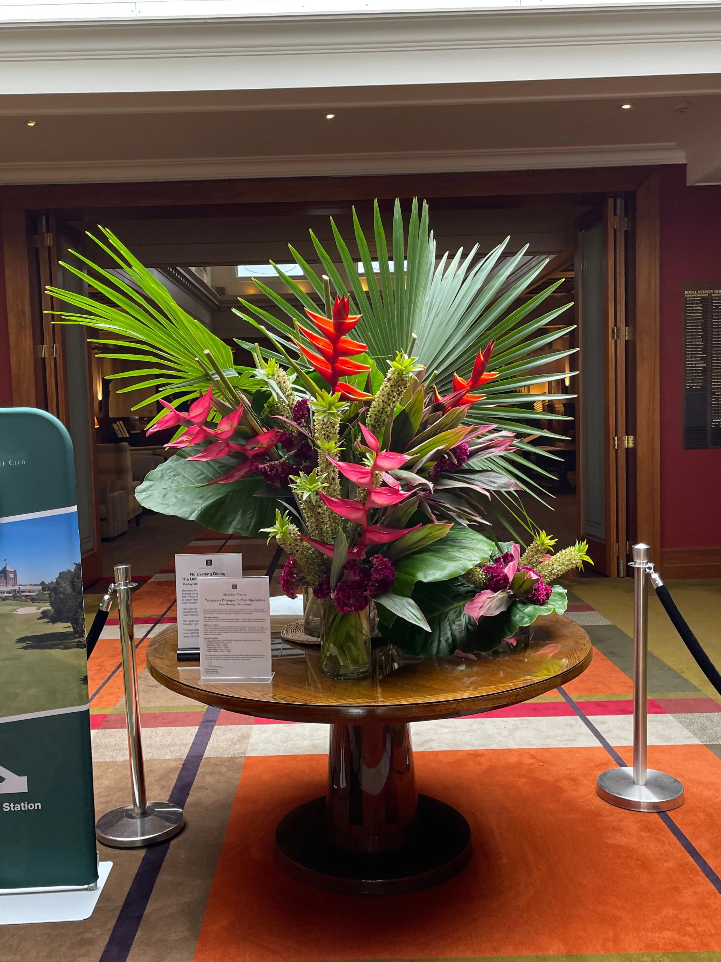 A large floral arrangement with tropical plants and colorful flowers on a round wooden table in an indoor setting with carpeted flooring and a sign with information.