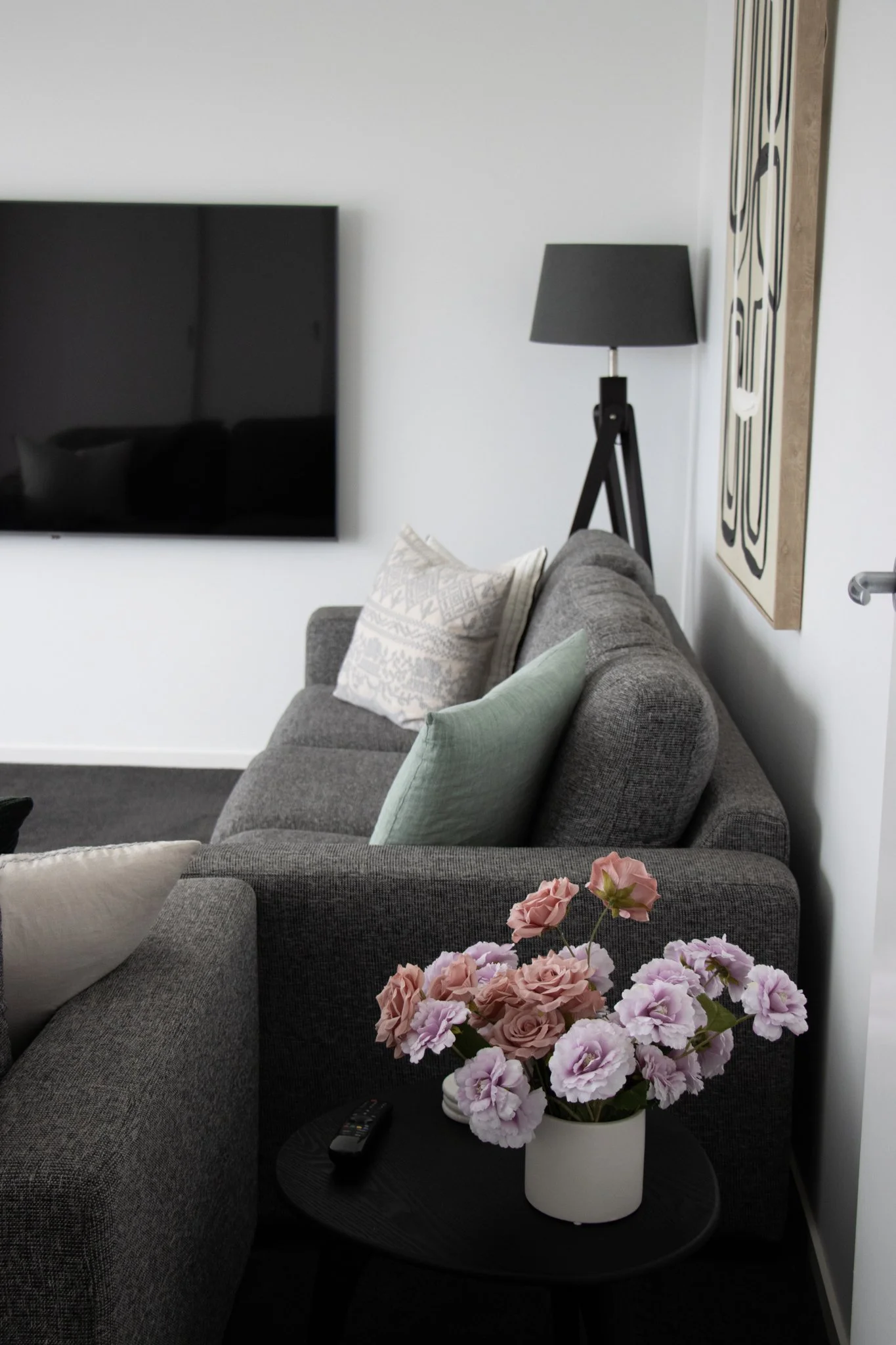 Living room with a gray sofa, a black side table with a pink and purple flower arrangement, and a flat-screen TV on the wall.