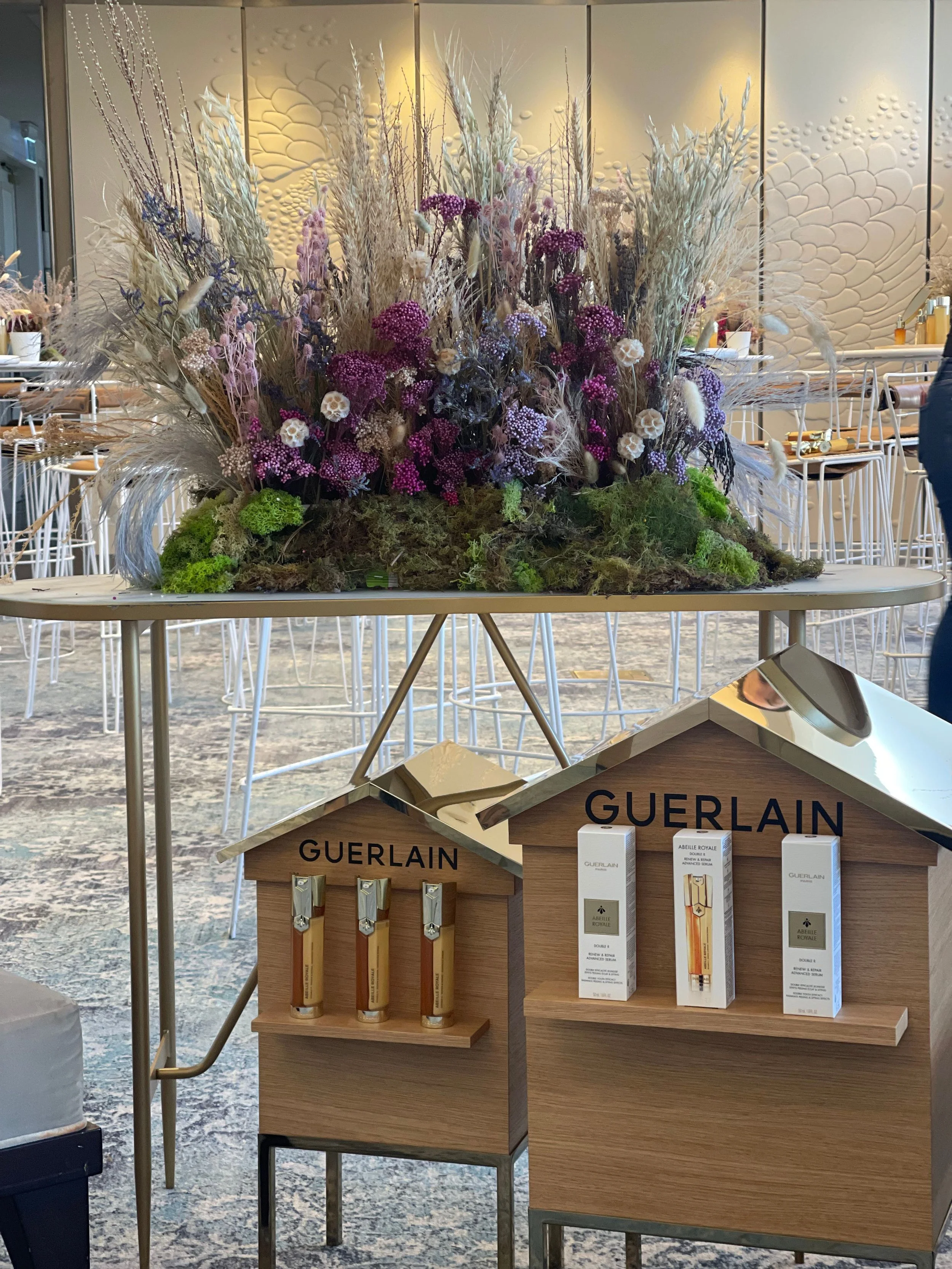 Display of Guerlain fragrance products and a floral arrangement on a table at an event or store.