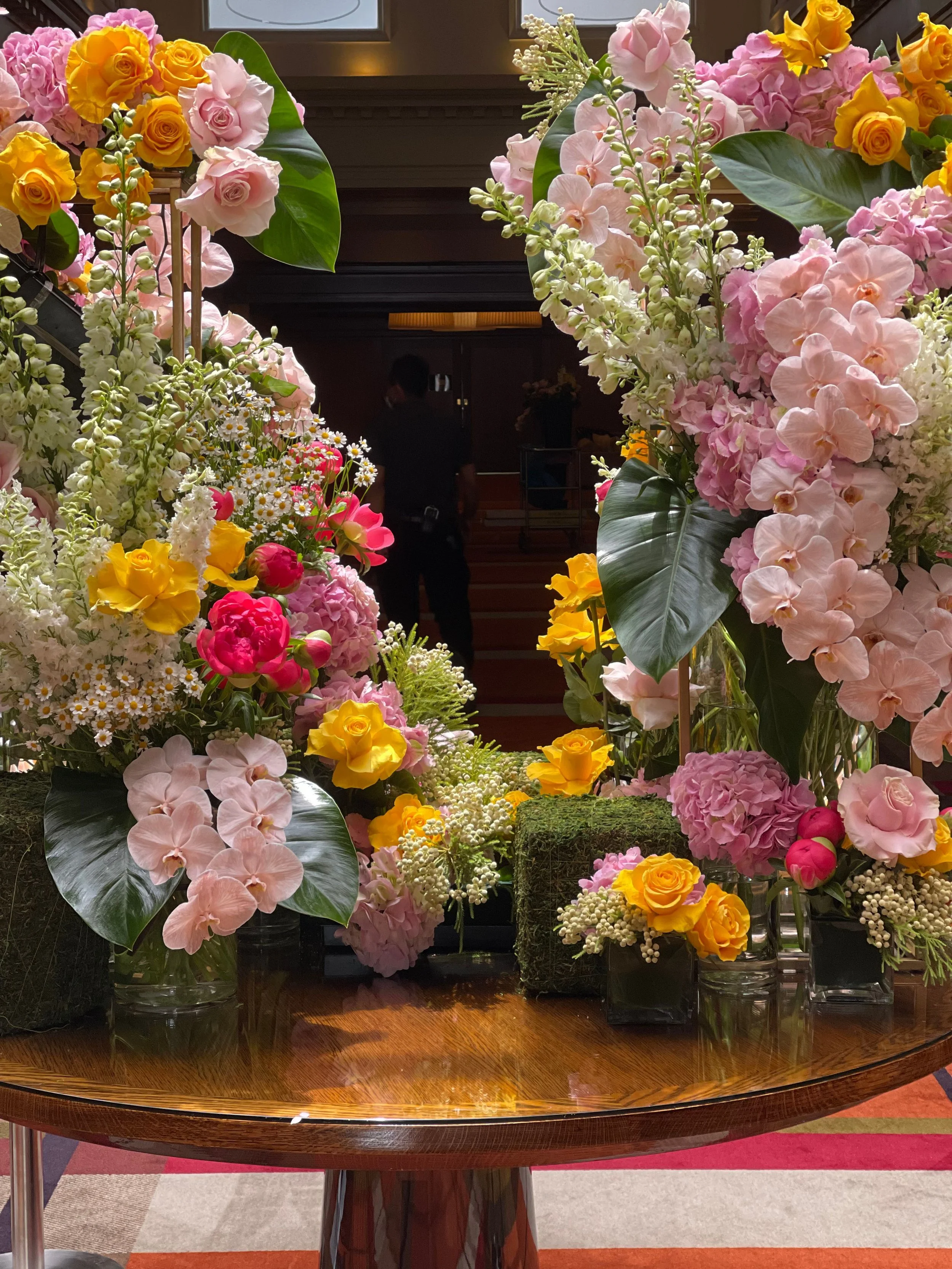 A table decorated with colorful flower arrangements including pink, yellow, and white flowers, with large green leaves, in an indoor setting.