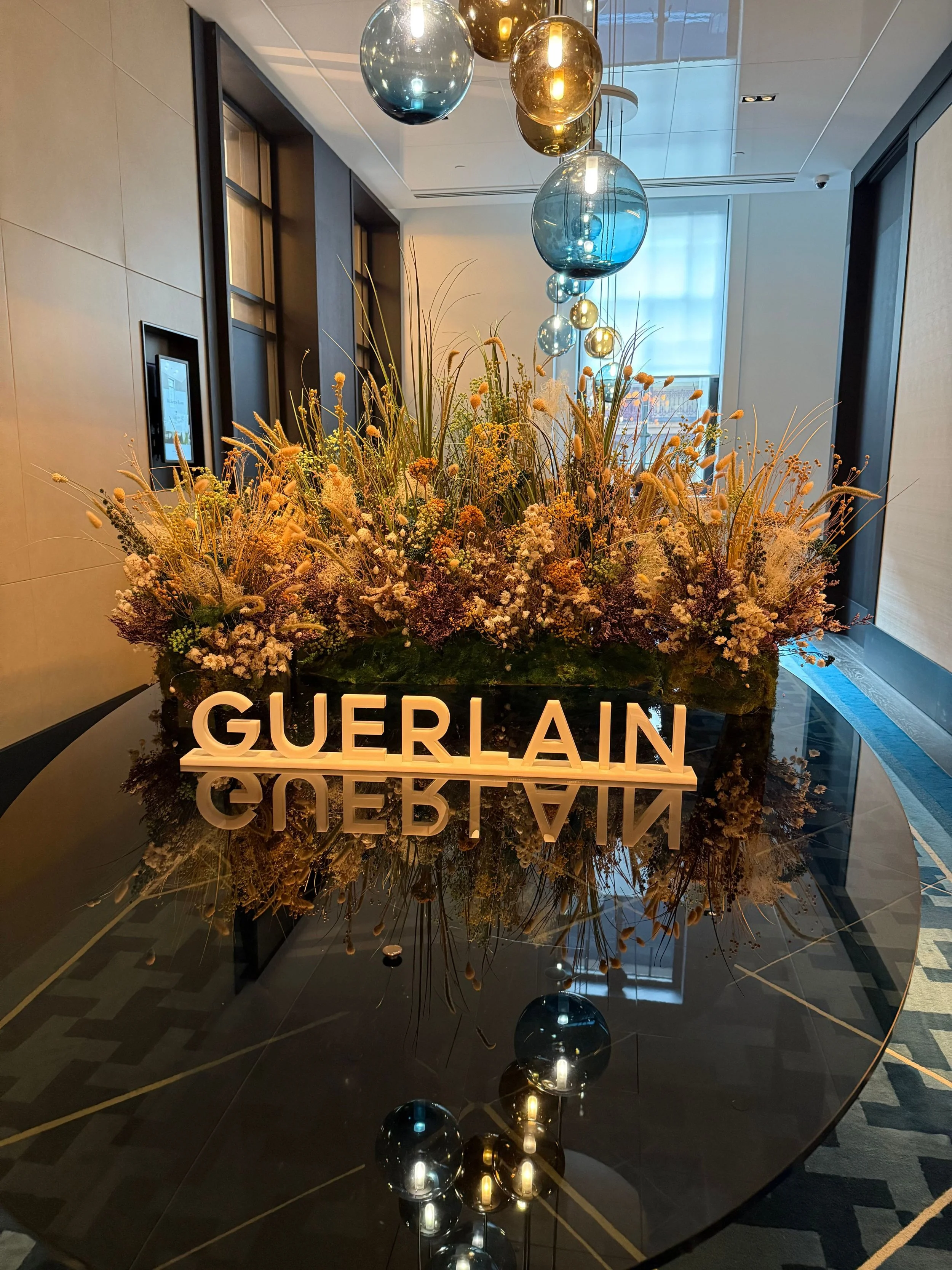 A black glossy table with a floral arrangement and a white 'GUERLAIN' sign. Above the table are hanging glass orbs in various shades of blue, gold, and clear. The background shows a modern interior with beige and dark-colored walls, a window with cur