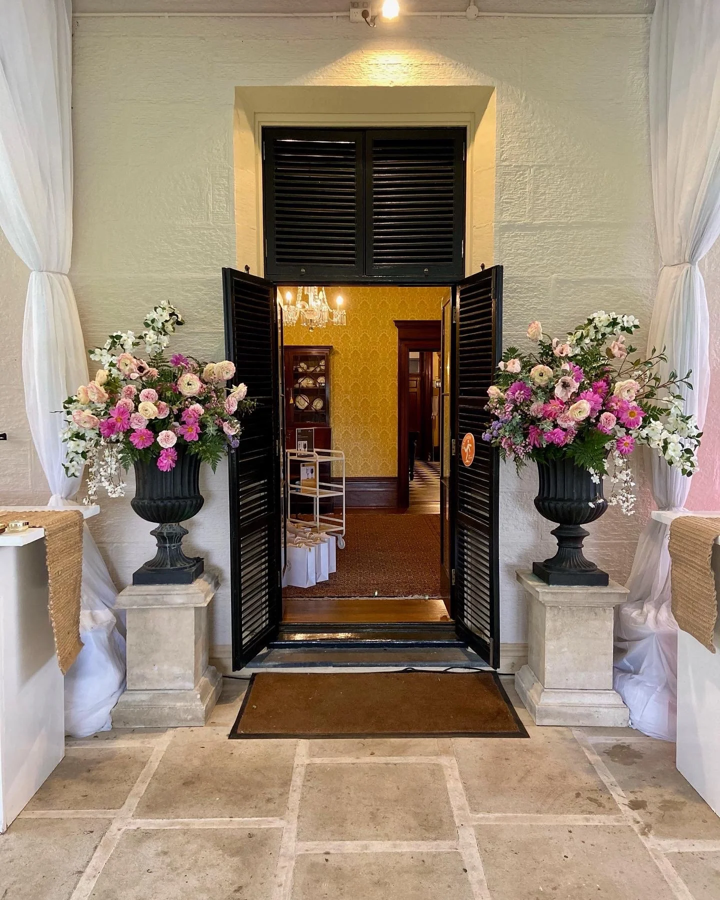 Open black shutters on a doorway with floral arrangements on stone pedestals on either side, leading into a warmly decorated interior with yellow wallpaper, a chandelier, and wooden furniture.