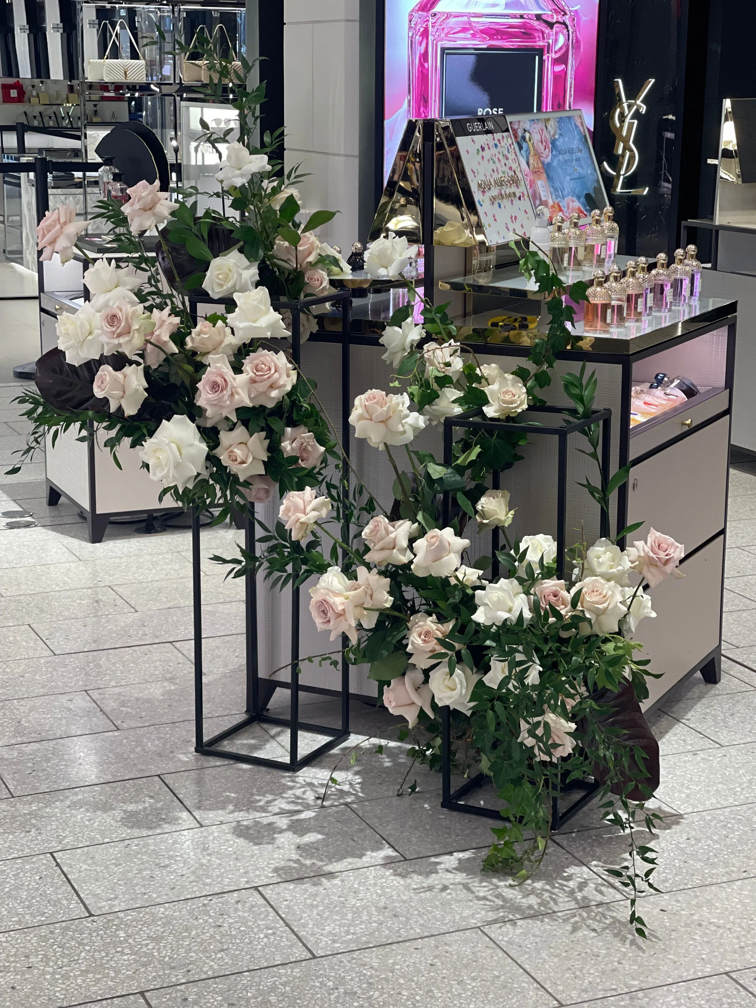 Display of perfumes and beauty products in a store, decorated with a large arrangement of white and light pink roses and green leaves.