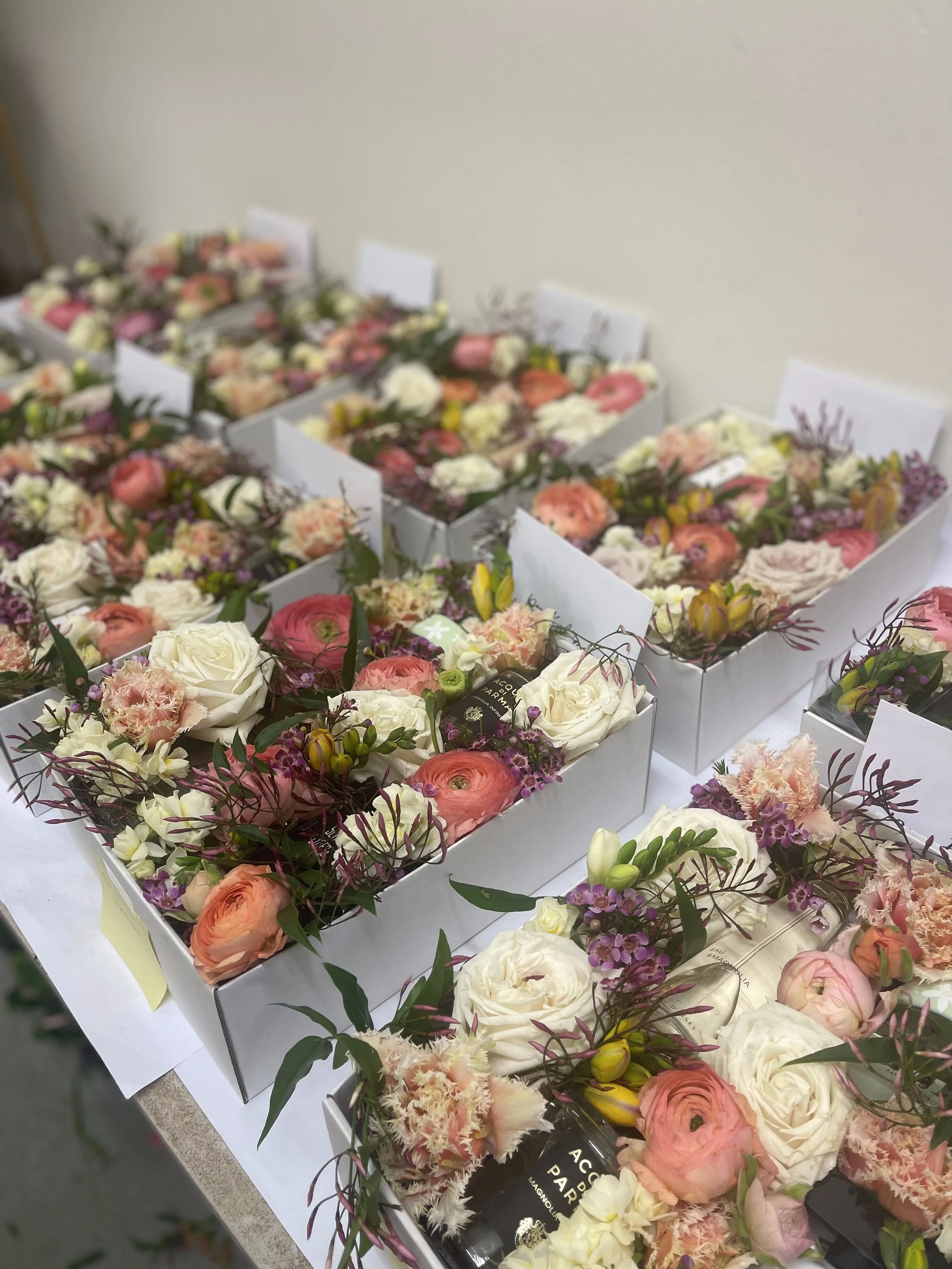 Multiple white boxes filled with various pink, white, and peach flowers, neatly arranged on a table, with some greenery and small decorative elements.