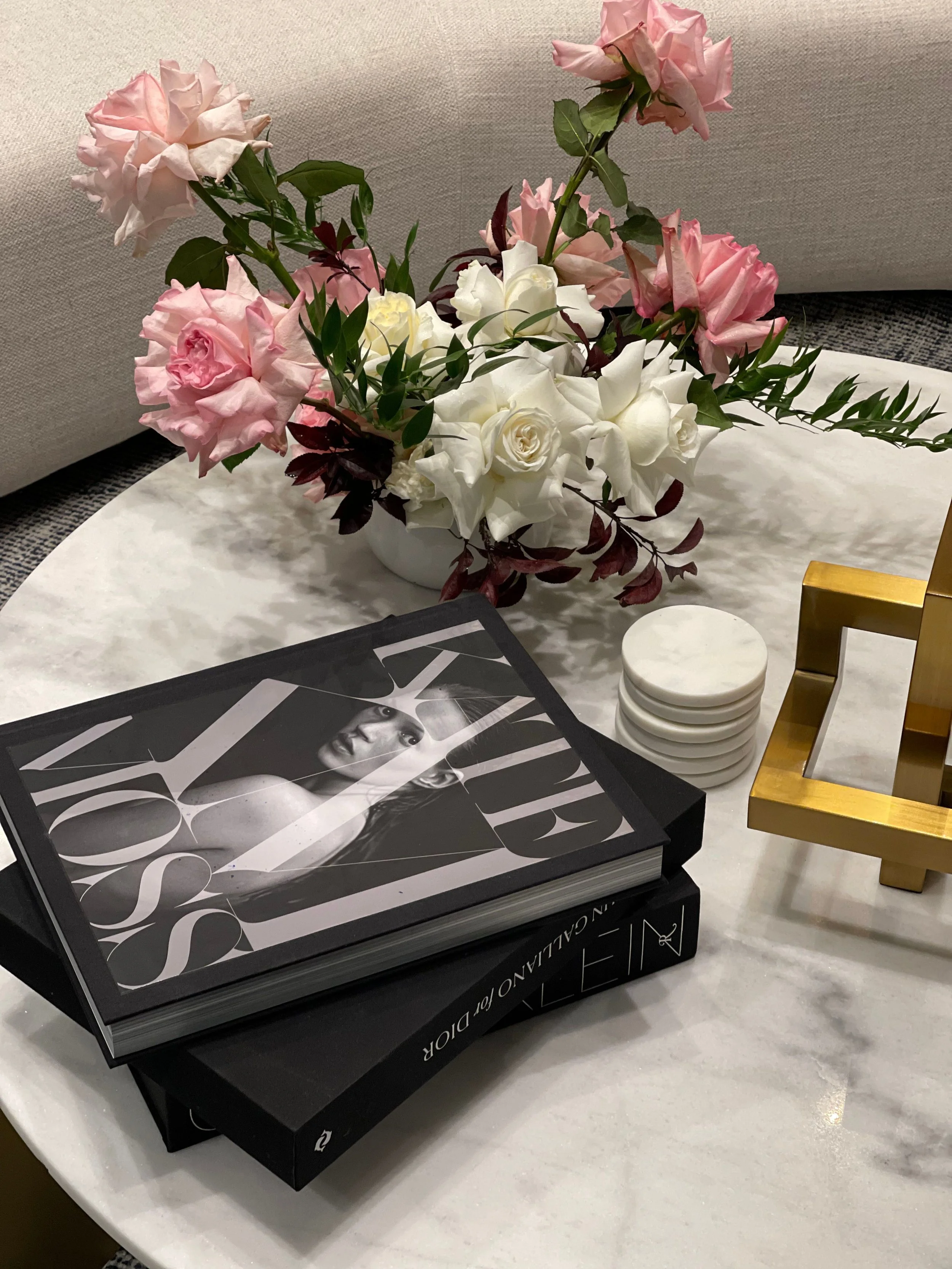 A marble table with a bouquet of pink, white, and dark red flowers in a white vase, a stack of white coasters, a large black and white fashion magazine on top of a black book, and a gold decorative object.