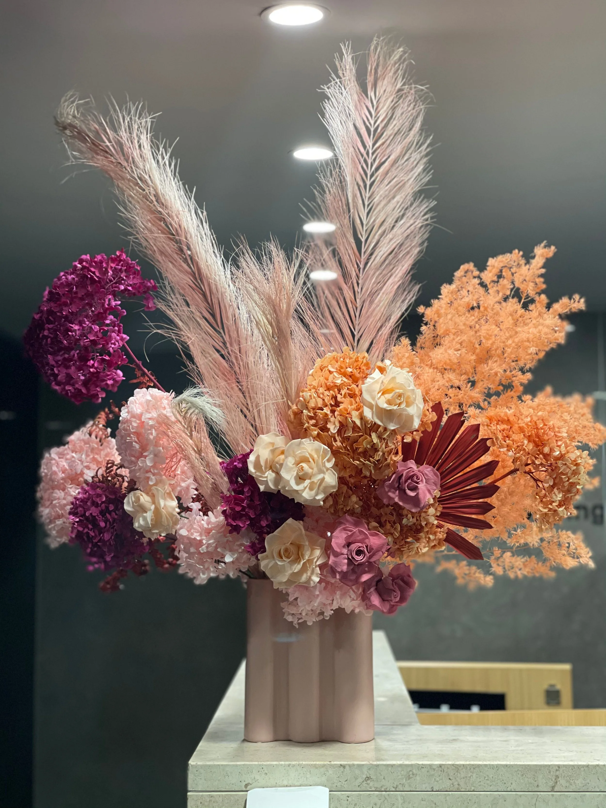 A large floral arrangement featuring pink, purple, cream, and orange flowers along with tall, wispy pink and beige pampas grass in a light pink vase on a countertop.