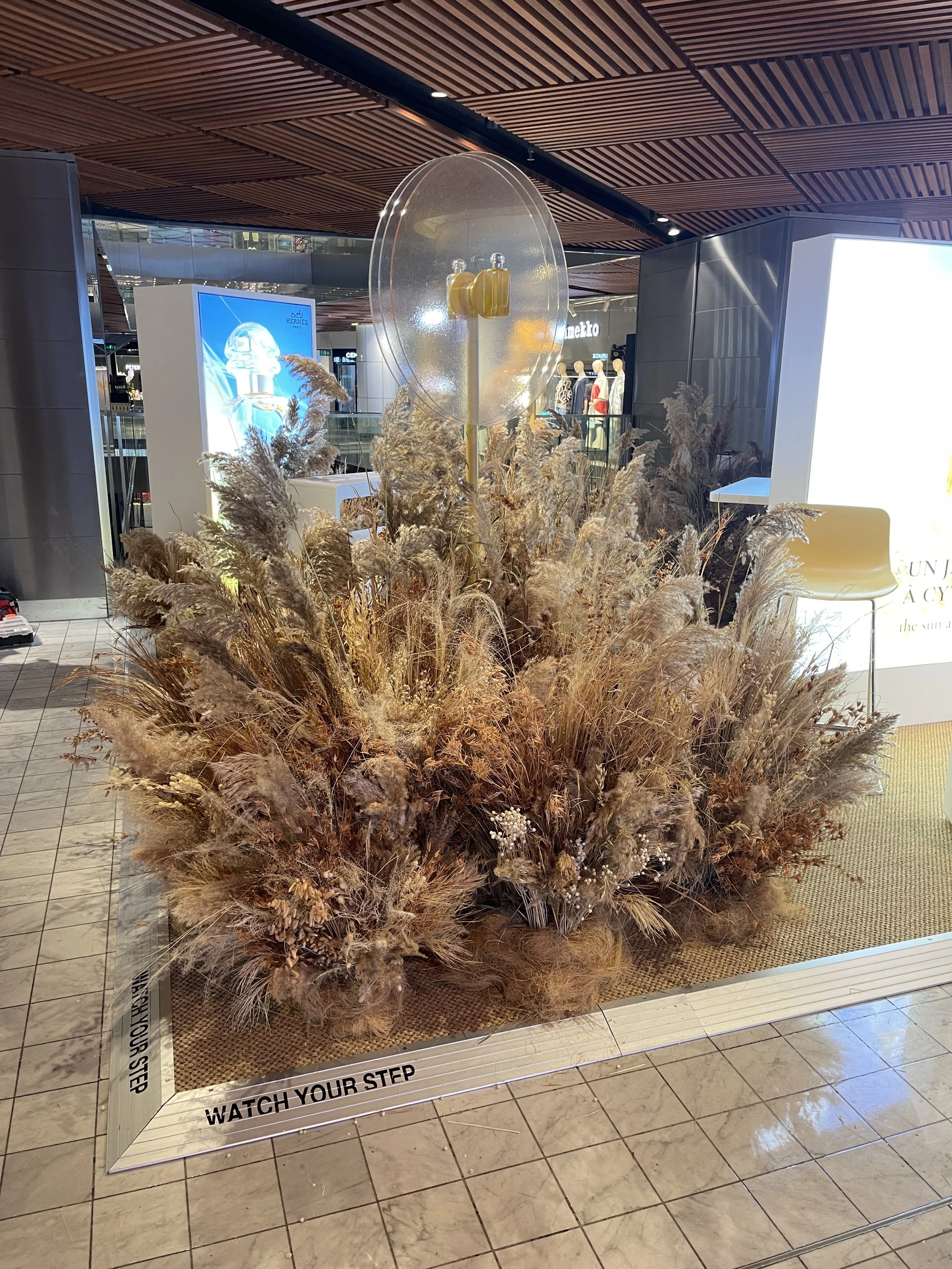 Decorative display of dried grass and flowers with a clear sphere on top, placed in a shopping mall.