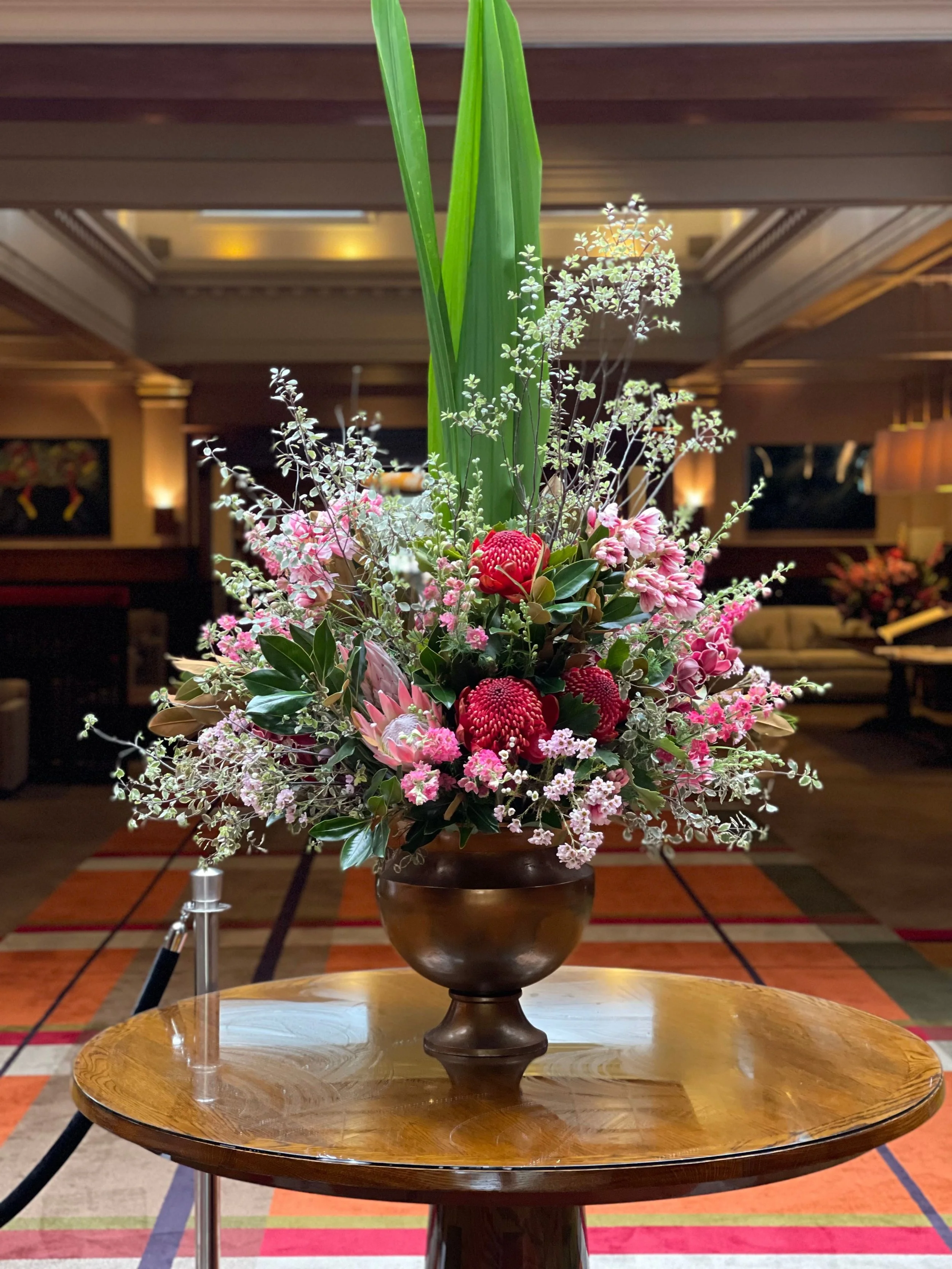 Elegant flower arrangement in a bronze vase on a wooden table, featuring pink, red, and white flowers with tall green leaves, in an indoor setting.