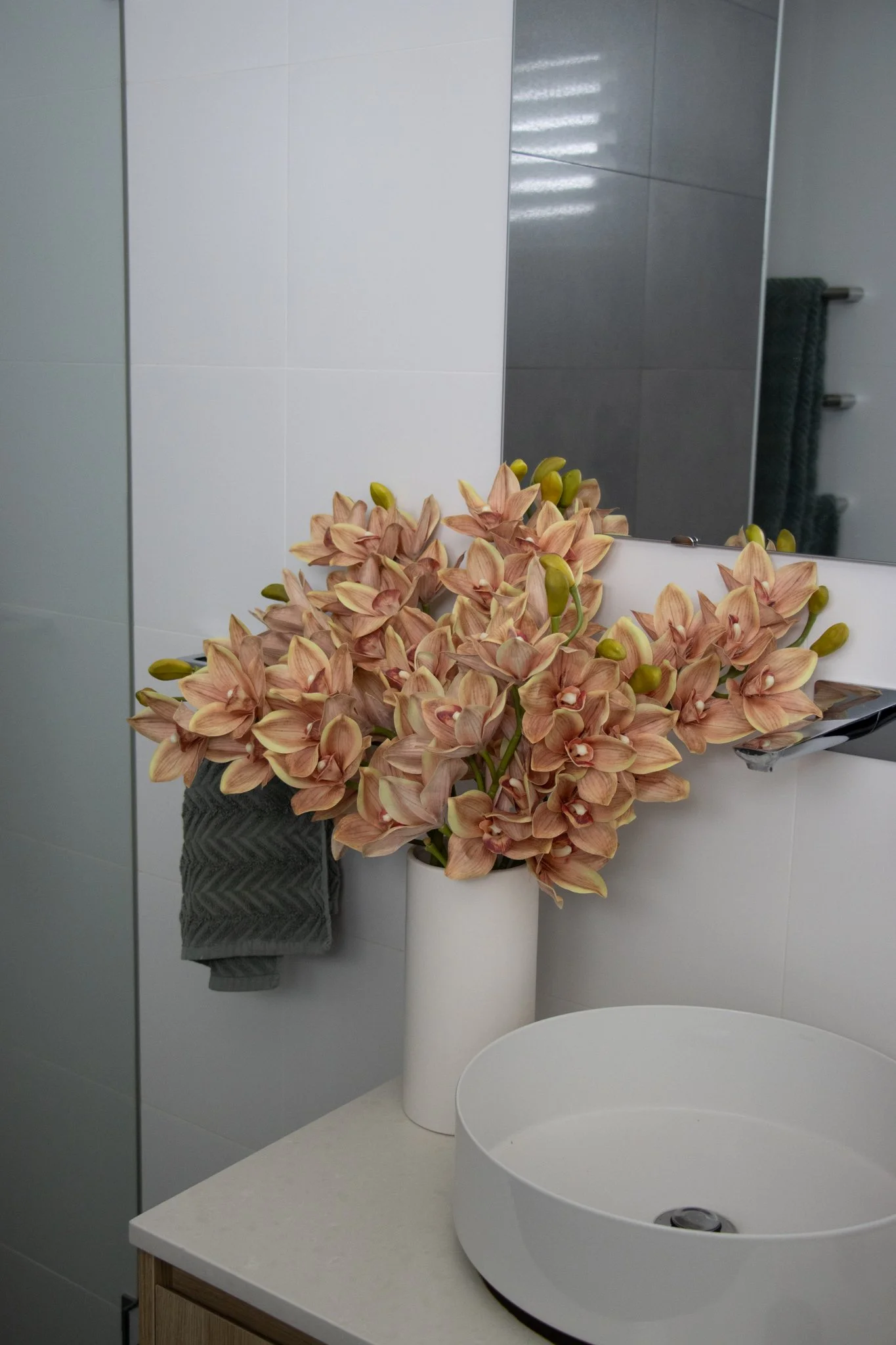 A bathroom sink area with a white round vessel sink, a white vase with pinkish-orange orchids, a gray hand towel, and a mirror reflecting part of the bathroom.