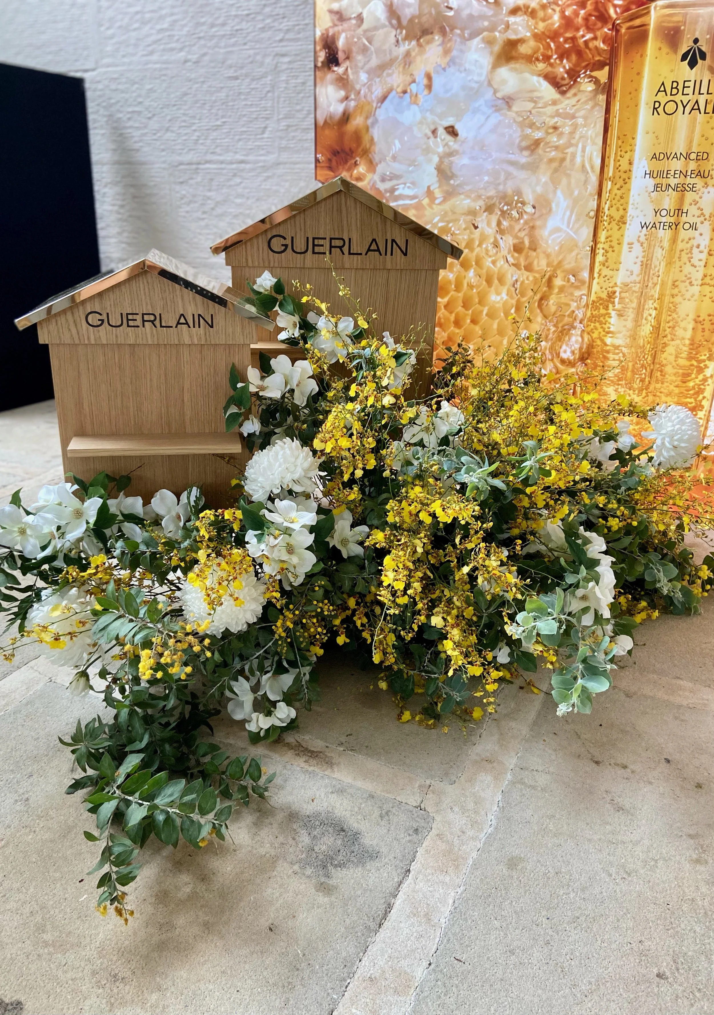 Decorative floral arrangement with white flowers and greenery, two wooden GUERLAIN display boxes, and a BEYONCE ROYAL product poster in the background.