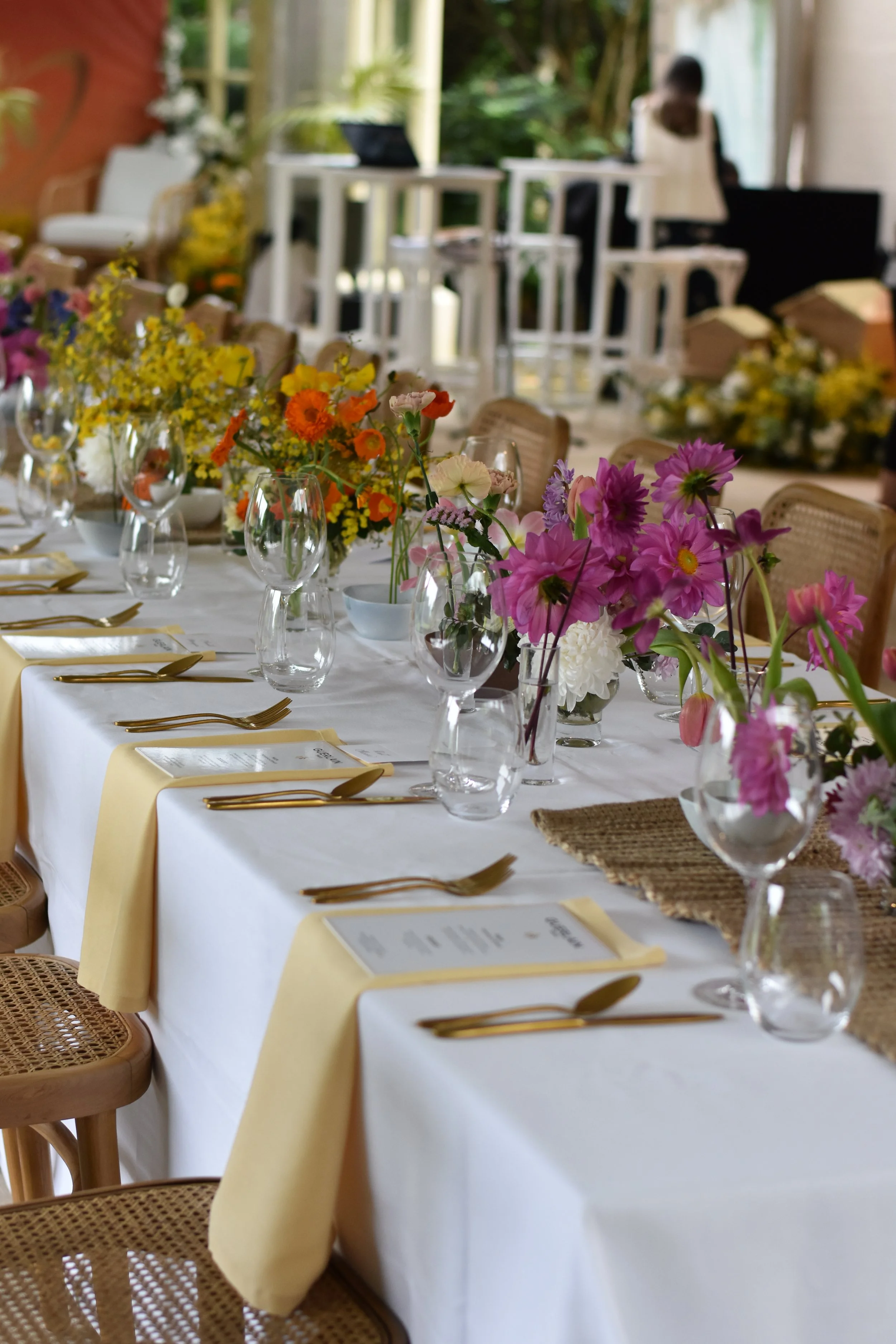 A long banquet table decorated with floral centerpieces and set with gold utensils, glasses, and menus, ready for a celebration or event.