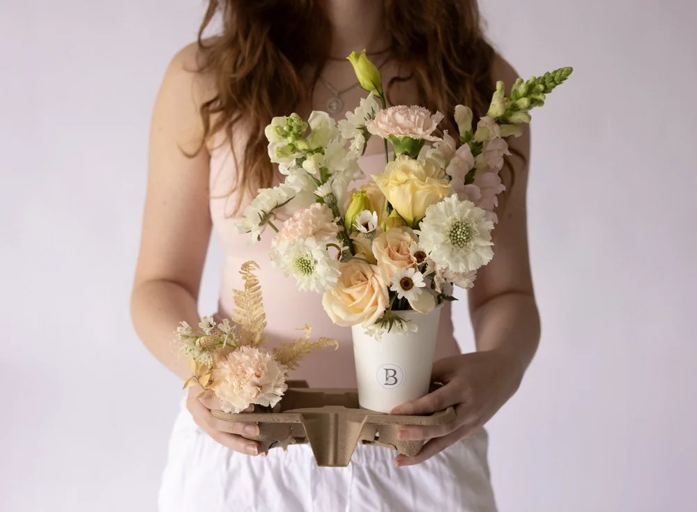A young girl in a white dress with floral headband sitting on grass, picking up flowers at an outdoor wedding setup with decorated chairs and large floral arrangements.
