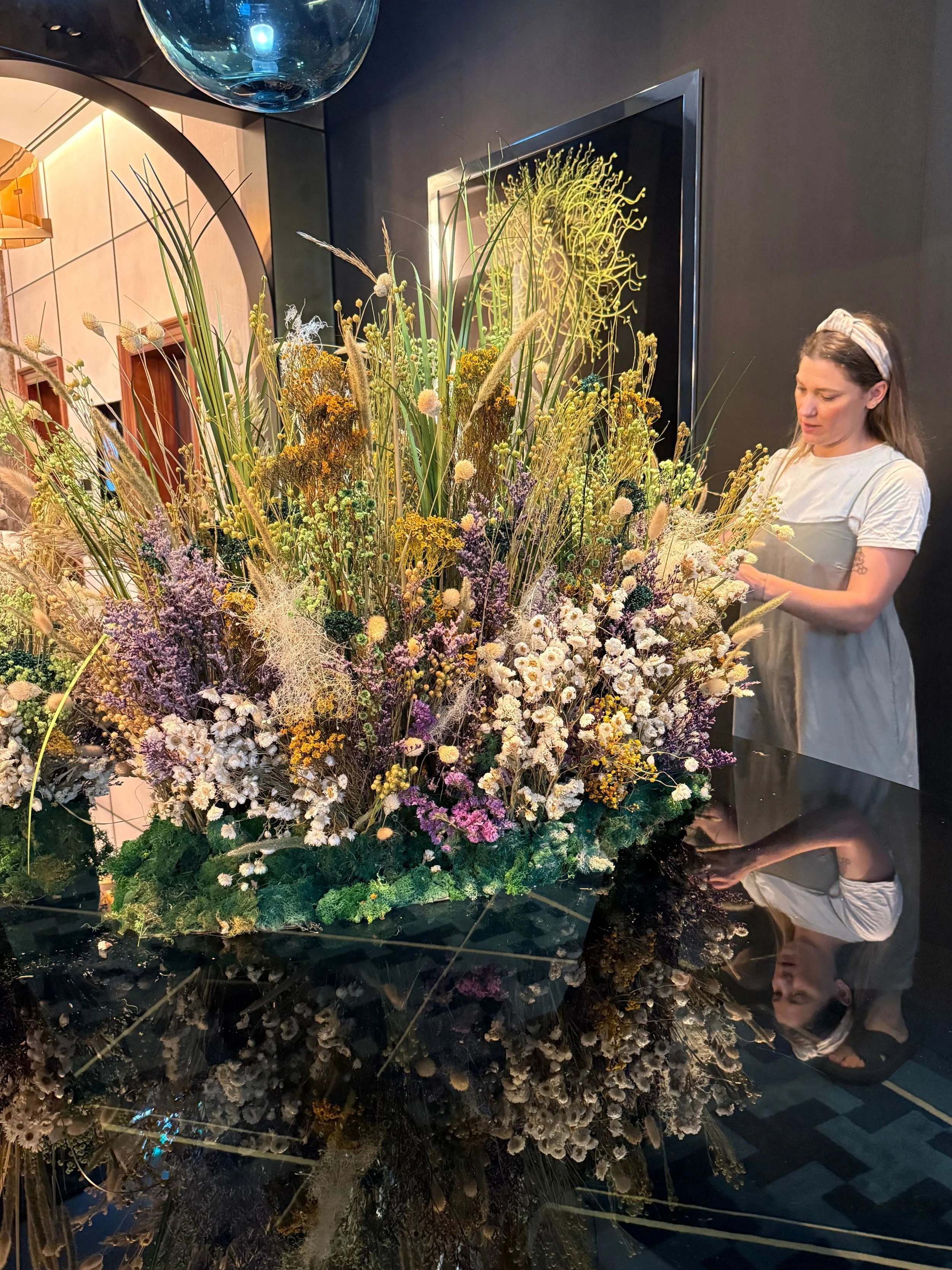A woman in a white and gray dress and headband arranging a large and colorful bouquet of flowers on a shiny black table in a modern interior setting.