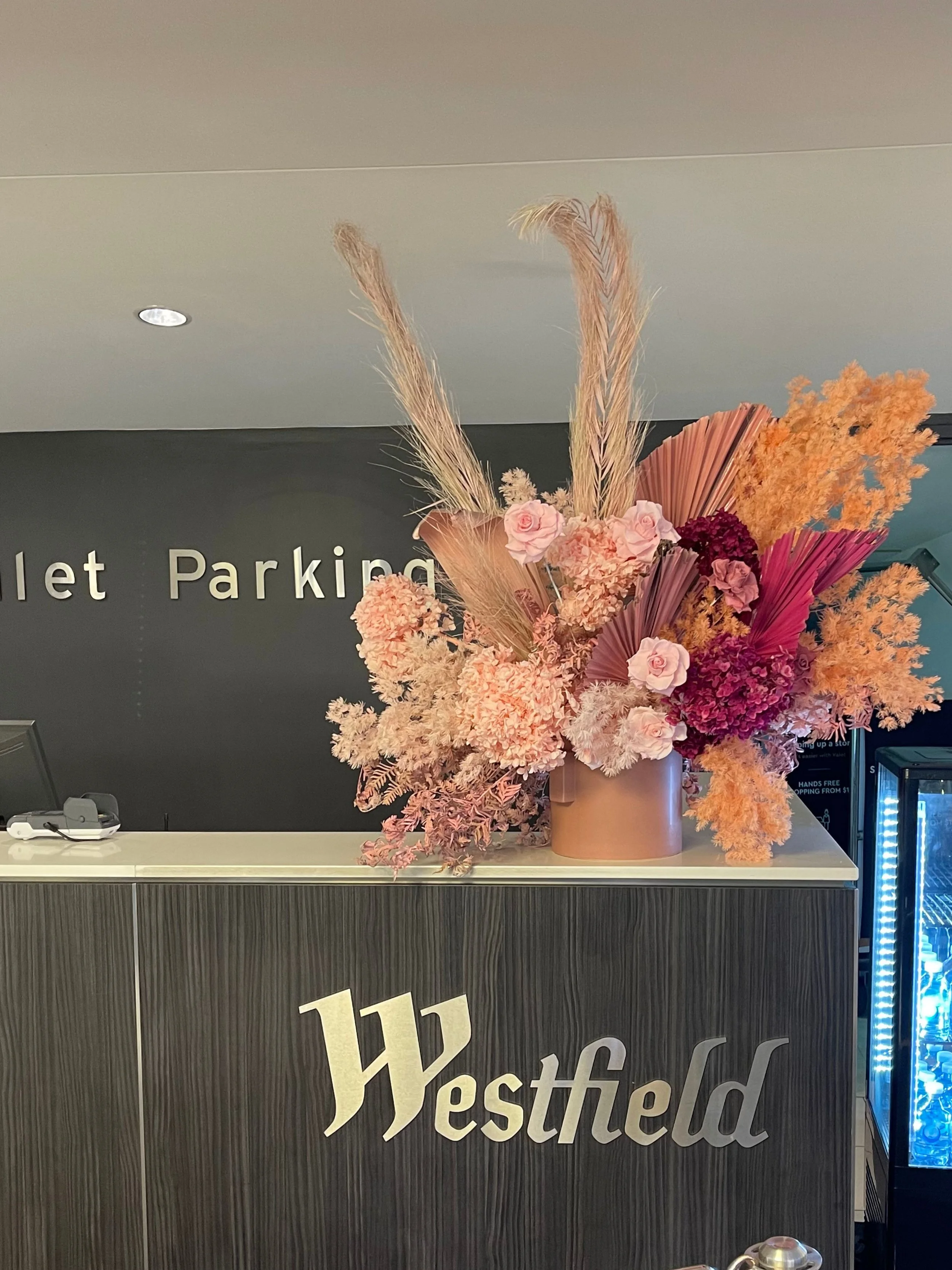 A large pink floral arrangement with pampas grass, roses, and dried leaves on a reception desk at Westfield mall.
