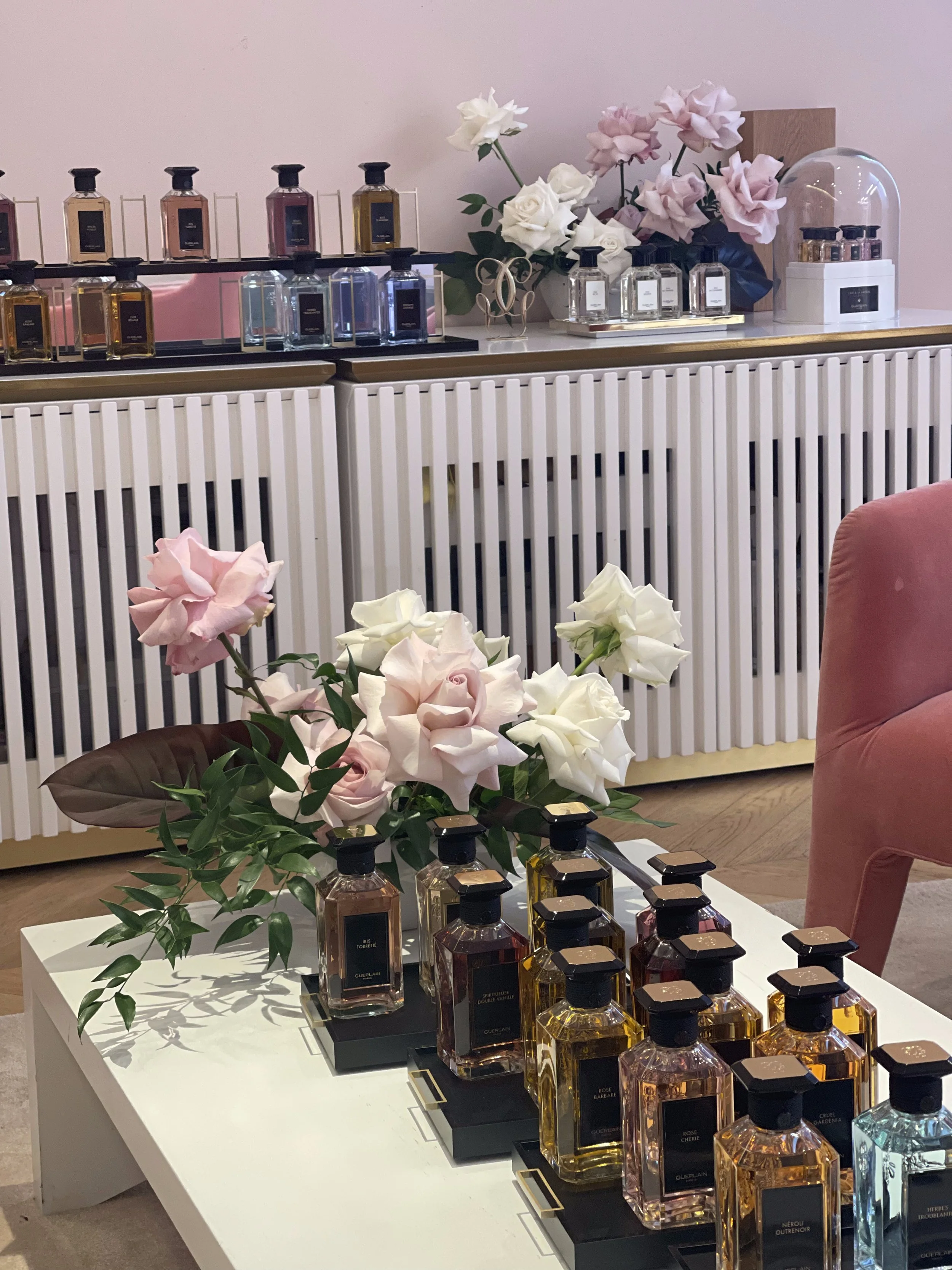 Display of perfume bottles and pink and white roses on a white table and a cabinet in the background.