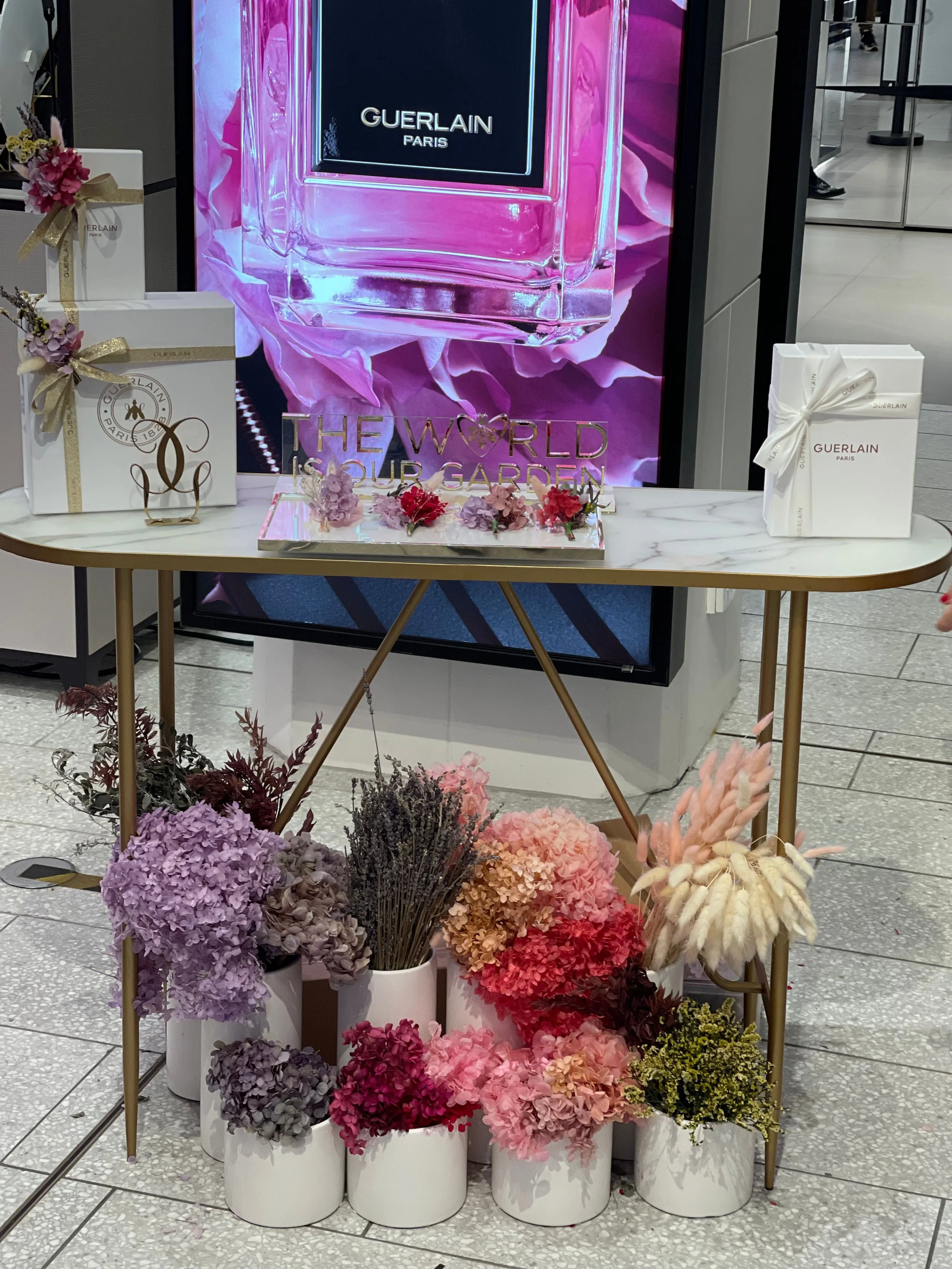 Display of luxury fragrances and floral arrangements at a Guerlain Paris promotional event, with a large digital screen in the background showing a perfume bottle and flowers.