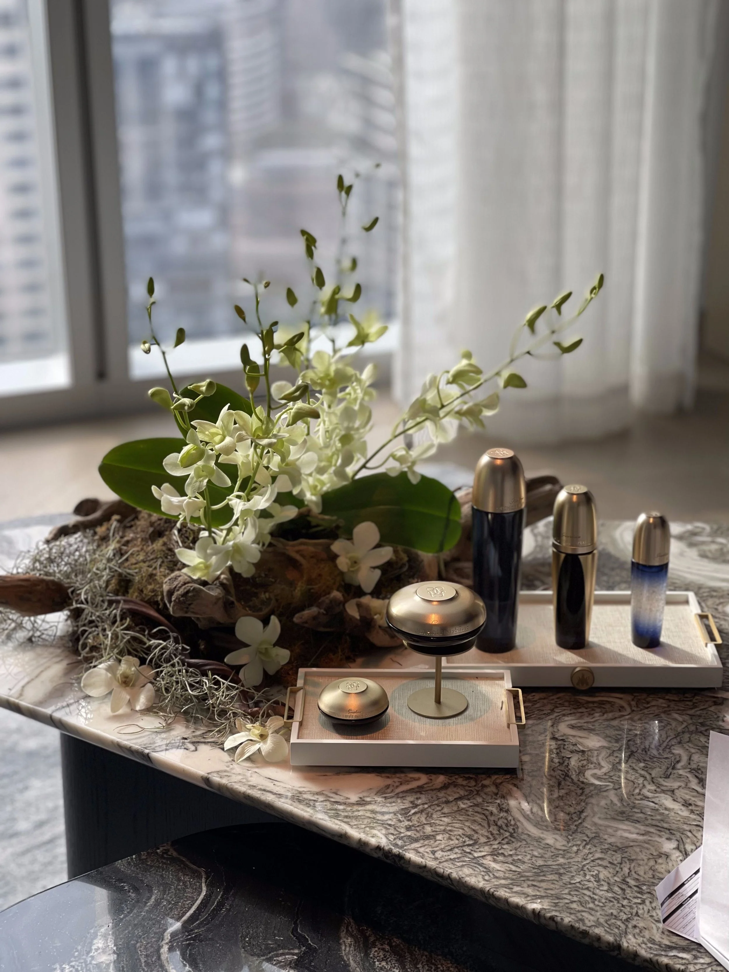 Decorative arrangement of white flowers and greenery on a marble table with perfume bottles and a tray, in front of a window with city buildings outside.
