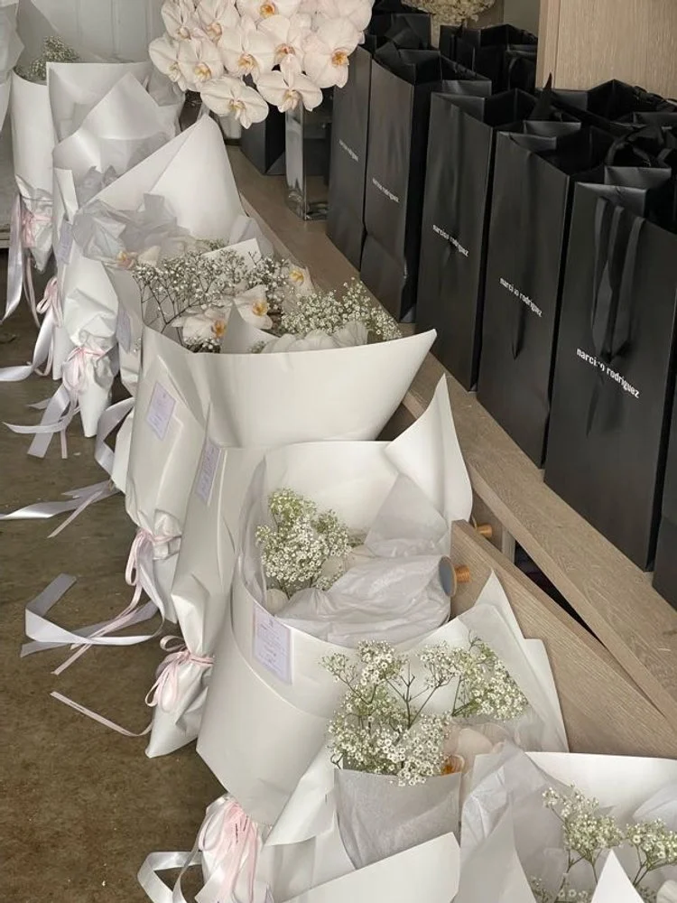 Various flower bouquets with baby's breath and white flowers wrapped in white paper and ribbons, arranged on a wooden surface with black shopping bags in the background.