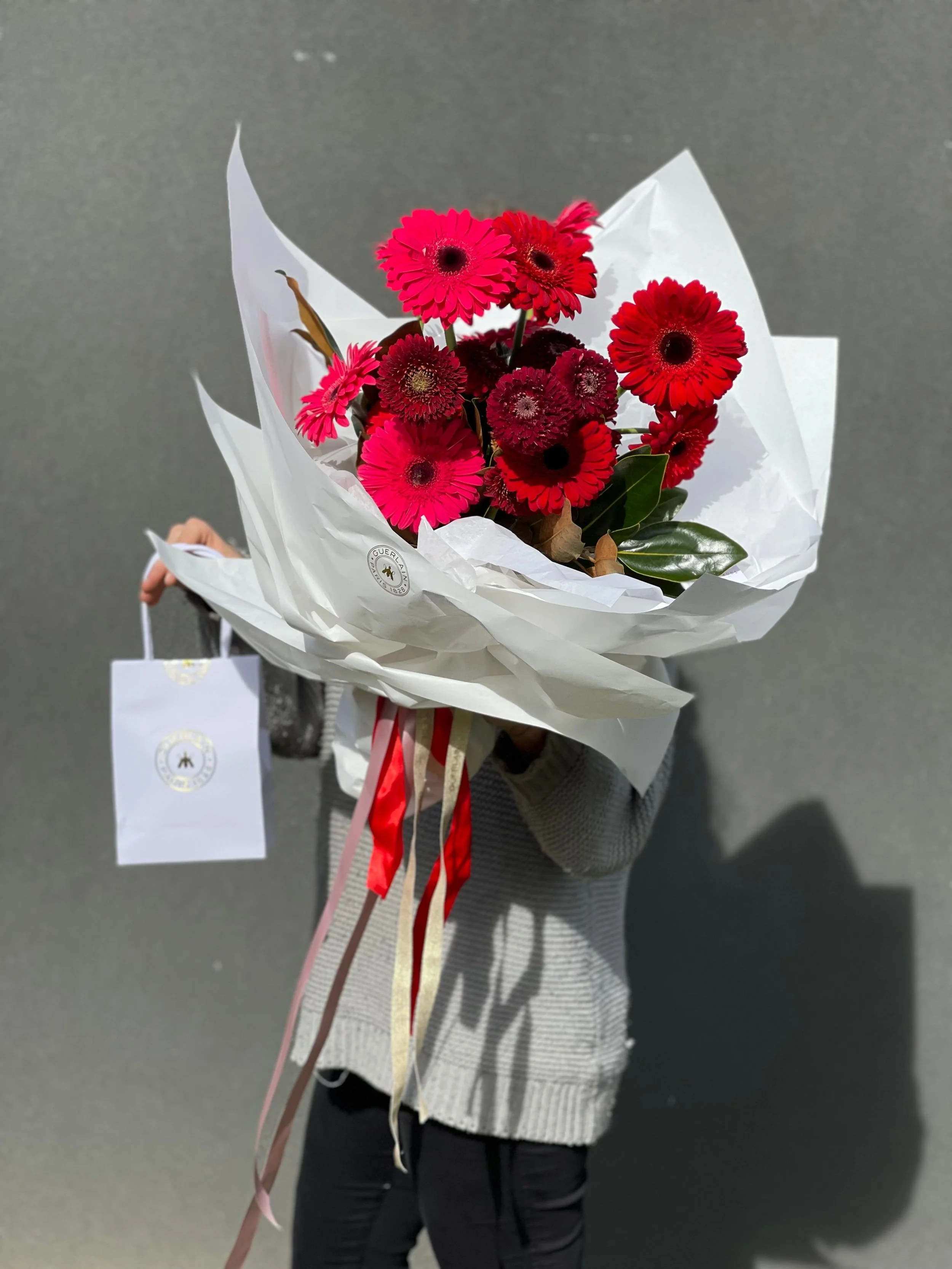 Person holding a bouquet of red and pink gerbera daisies wrapped in white paper with ribbons, against a gray background.