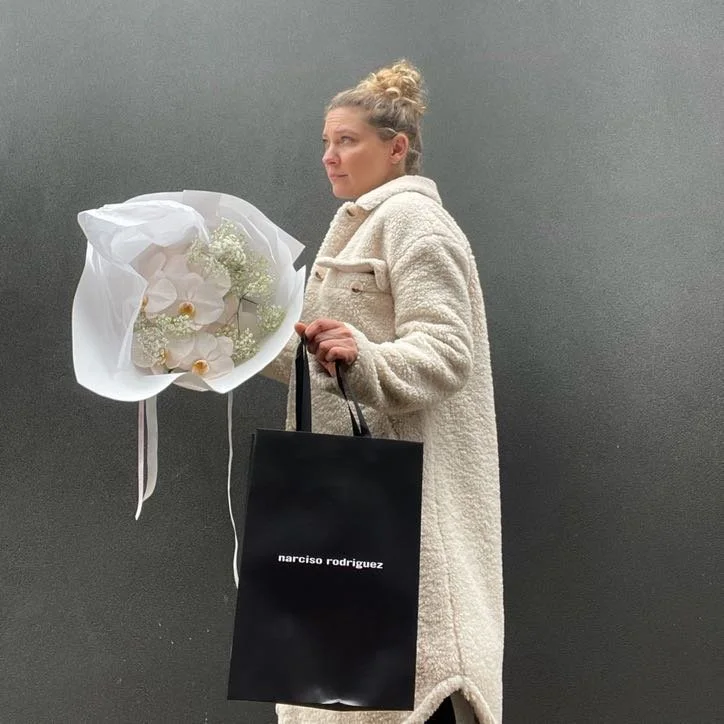 A woman with curly hair in a cream-colored coat holding a bouquet of white flowers and a black shopping bag that says 'narciso rodriguez' against a plain gray background.