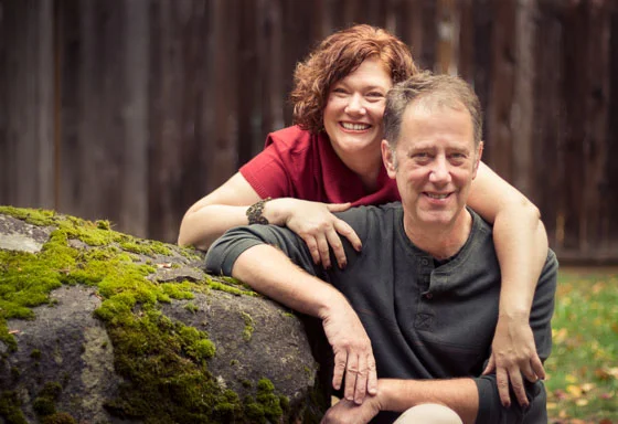 Couple smiling at the camera with arms draped over the other outside by a large mossy rock.