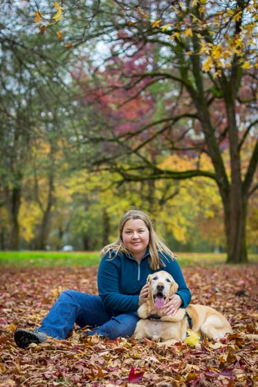 Person in blue sitting in fall leaves with yellow lab dog.