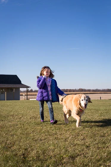 Child in purple coat with dog running in yard on a sunny day.