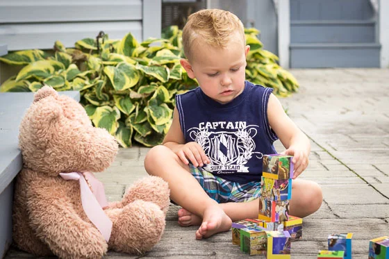 Small child in blue shirt outside playing with blocks and teddy bear.