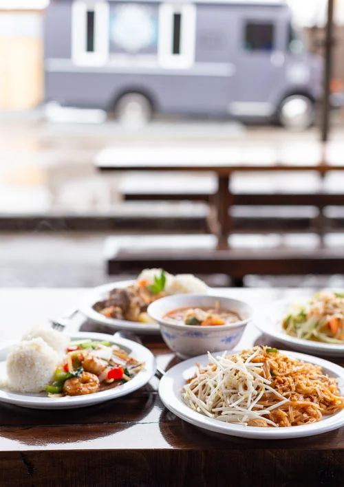 Table of Thai food dishes with picnic tables and food cart in background
