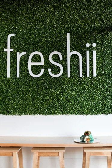 Wall of greenery with white Freshii lettering above a table and stools