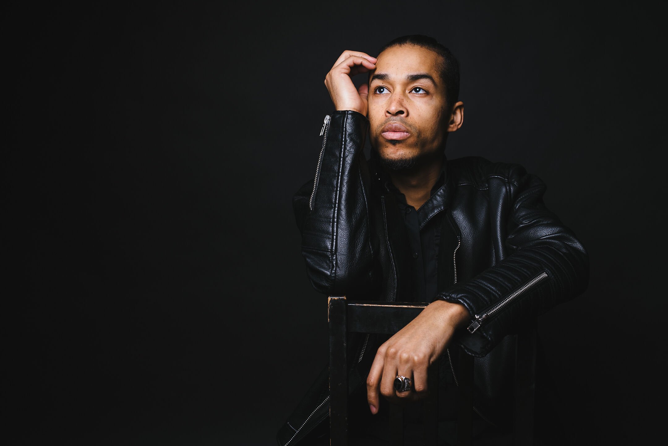 Portrait of a young man with braided hair, wearing a black leather jacket, sitting on a black chair against a black background, with a contemplative expression and hand resting on his head.