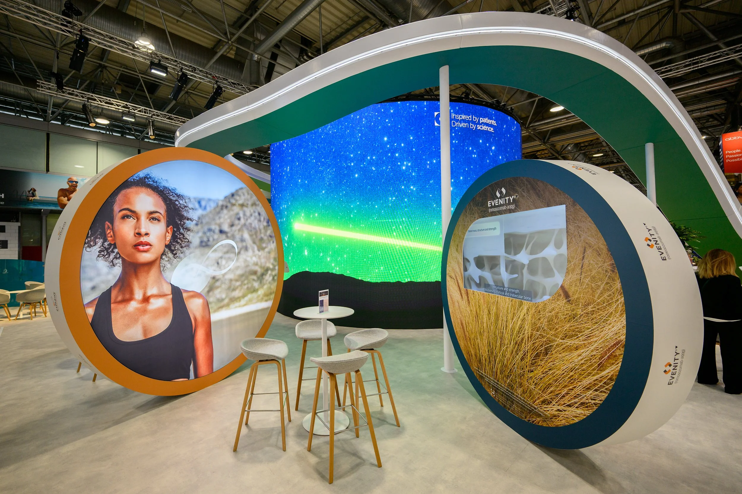Exhibition booth with large circular displays, one showing a woman with curly hair in a mountainous landscape, the other displaying dry grass. A backdrop features a starry night sky and green laser light.