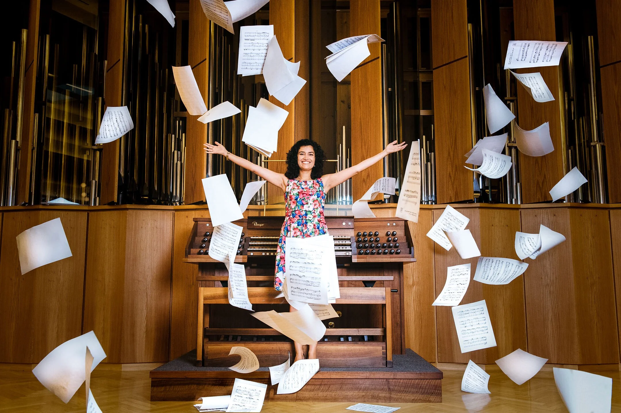 Woman in colorful dress standing on organ bench, smiling with arms outstretched, surrounded by flying sheets of music in a wooden music hall.