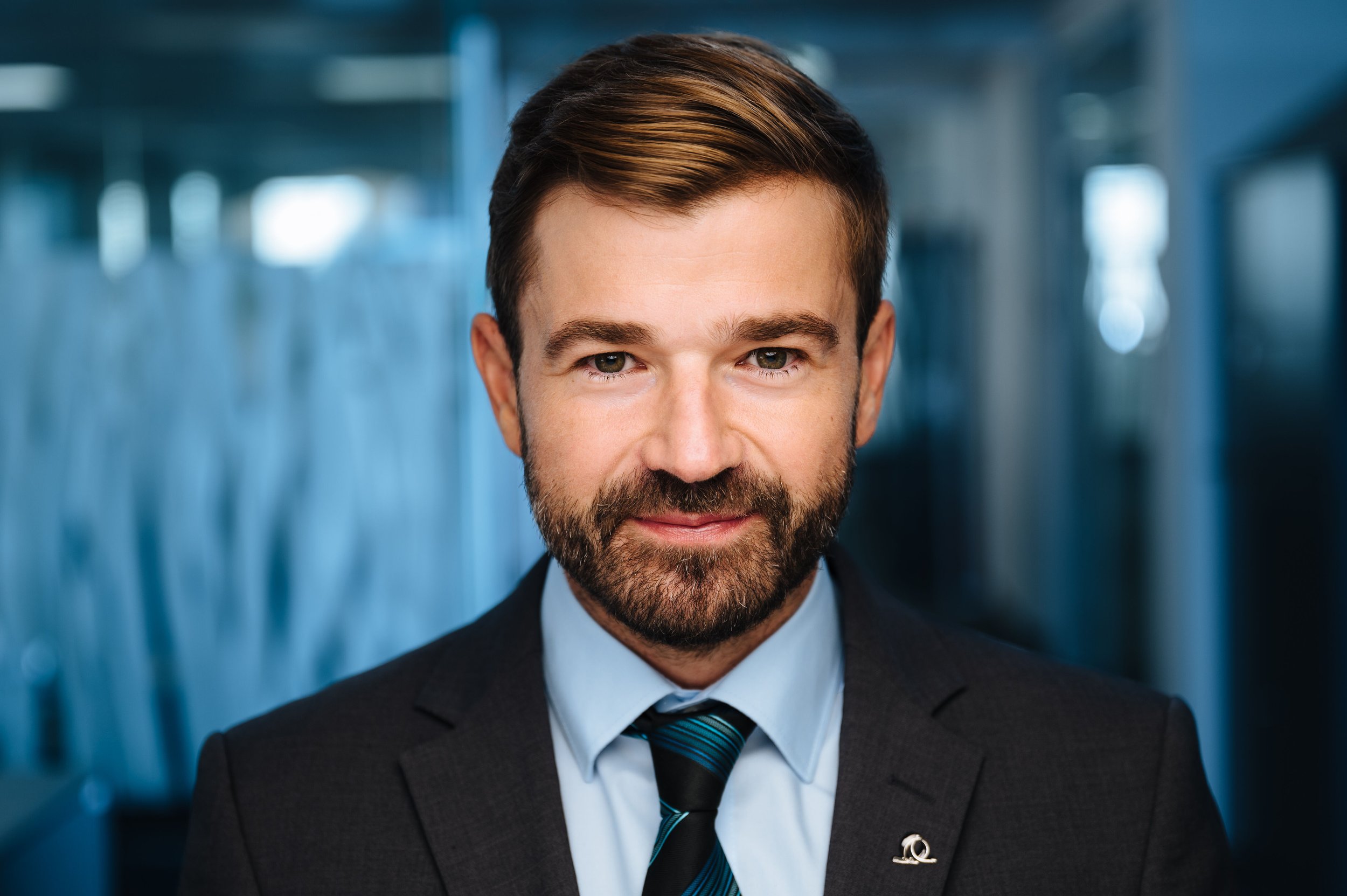 A professional man with brown hair, beard, and mustache in a suit and tie, standing in a modern office environment with blurred background.