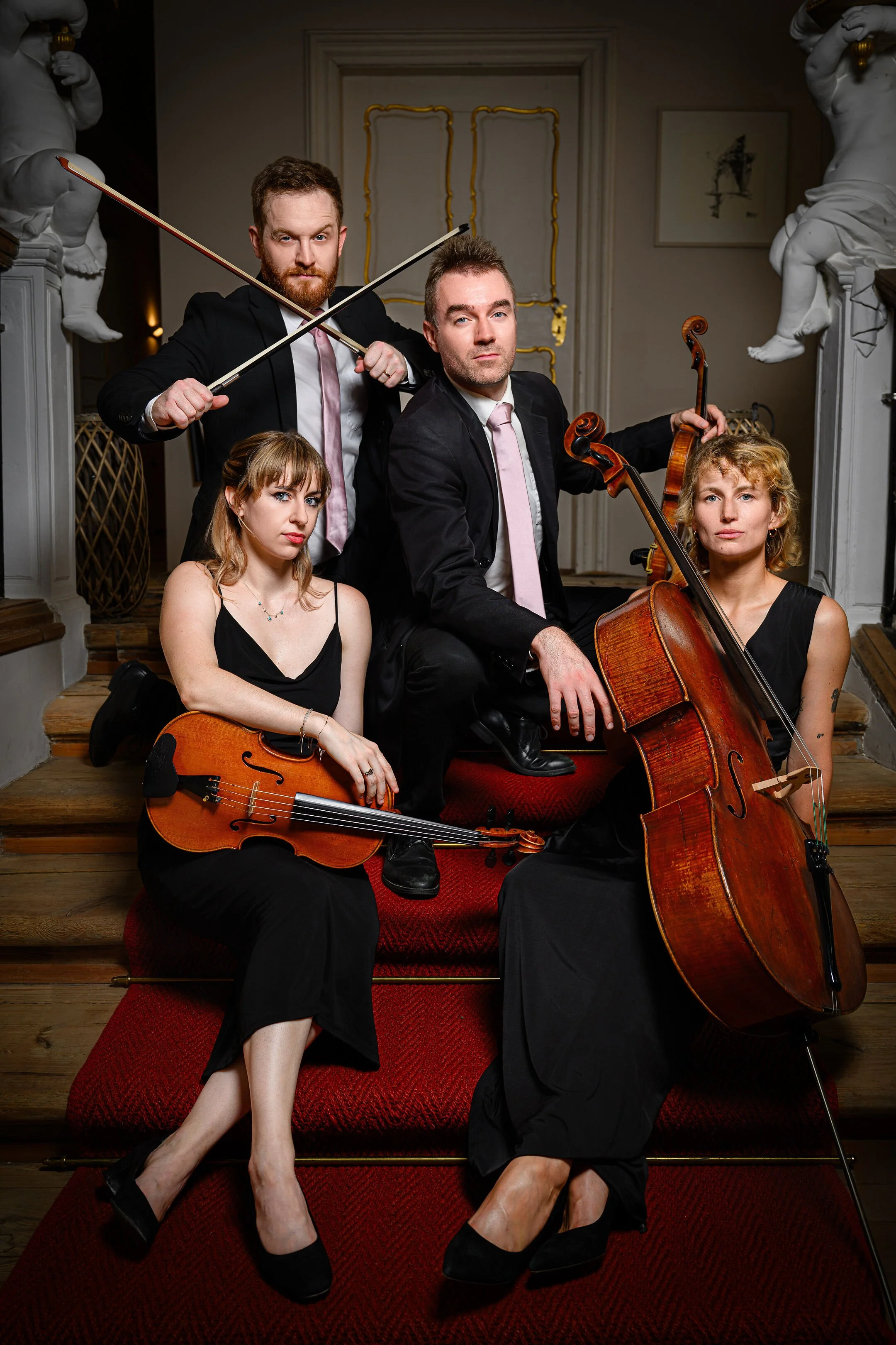 A group of four musicians in formal attire, with two women holding violins and a man holding a cello, and two men holding bows, posing on stairs inside an elegant room.