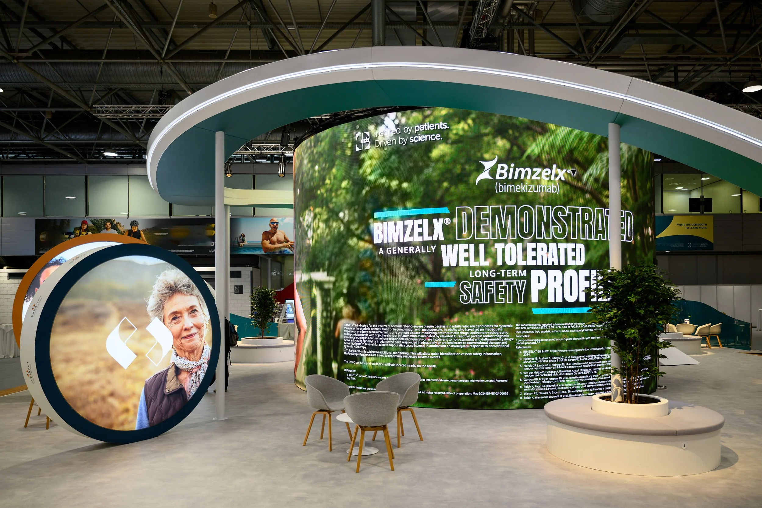 Trade show exhibit for BIMZELX (bimekizumab) featuring large digital display with information about the drug, a round digital screen with a woman's portrait, and seating areas with chairs and potted plants.