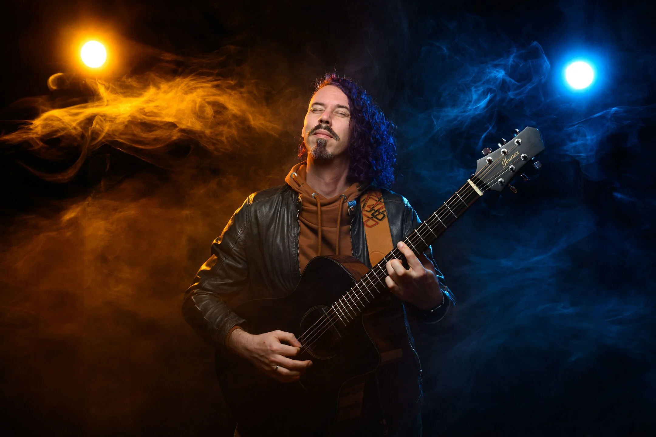 A man with long curly hair and a goatee playing an acoustic guitar with closed eyes, wearing a leather jacket, brown hoodie, and a guitar strap, surrounded by yellow and blue stage lights with smoky effects.