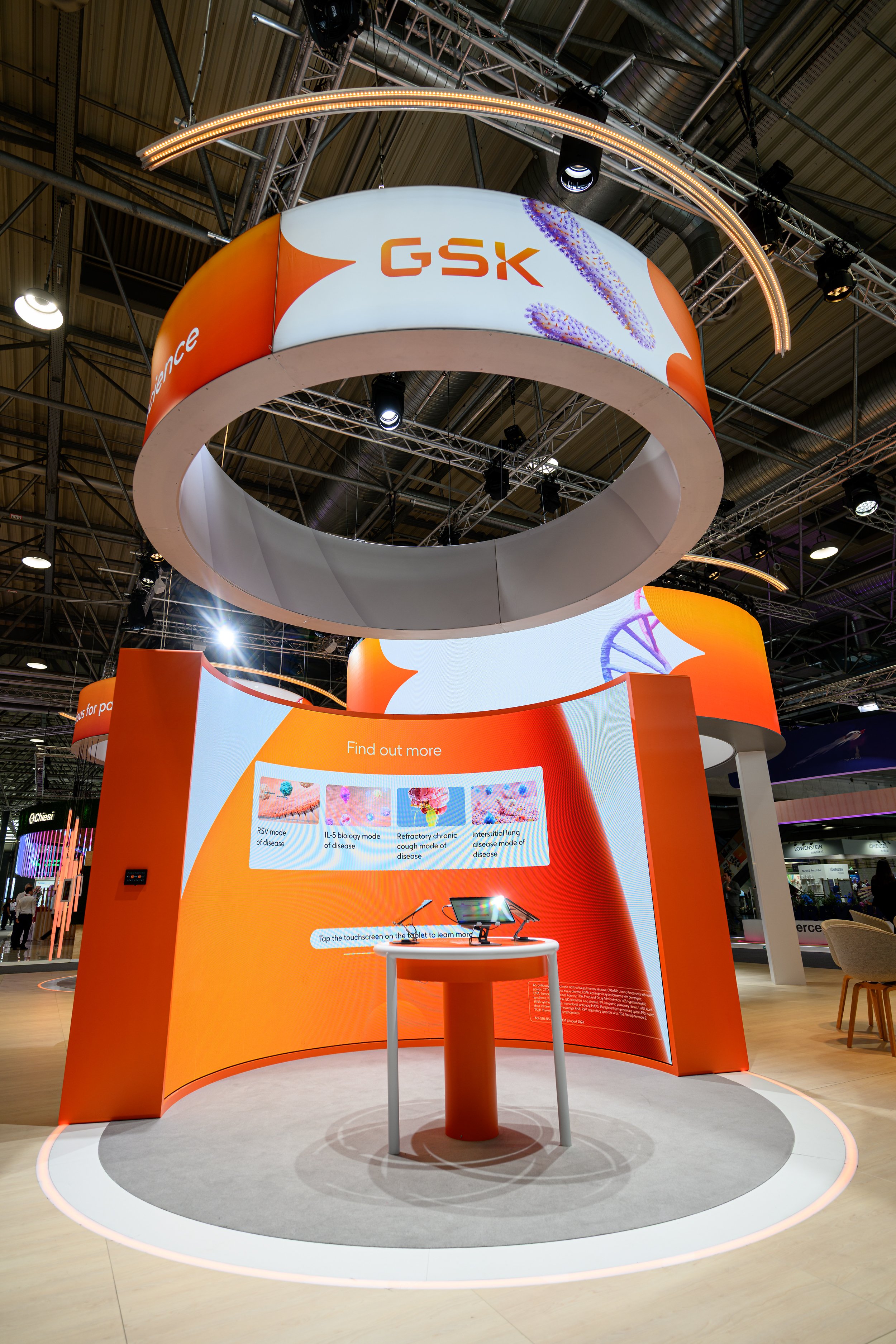 Exhibition booth featuring a large GSK sign with medical and scientific graphics, including DNA and disease images, on a circular orange and white display at a trade show or conference.