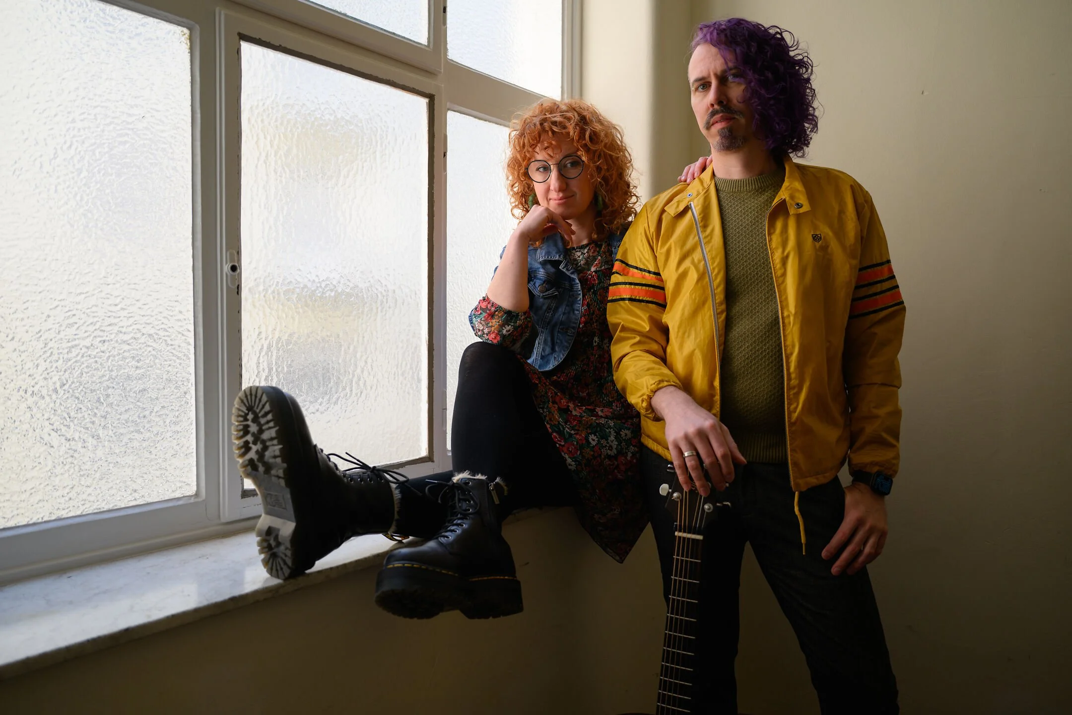 A duo of two adults stands next to a frosted window with textured glass. The woman with curly red hair and glasses sits on the window sill, wearing a floral dress and a denim jacket, and has her leg extended with a black boot. The man with curly purp