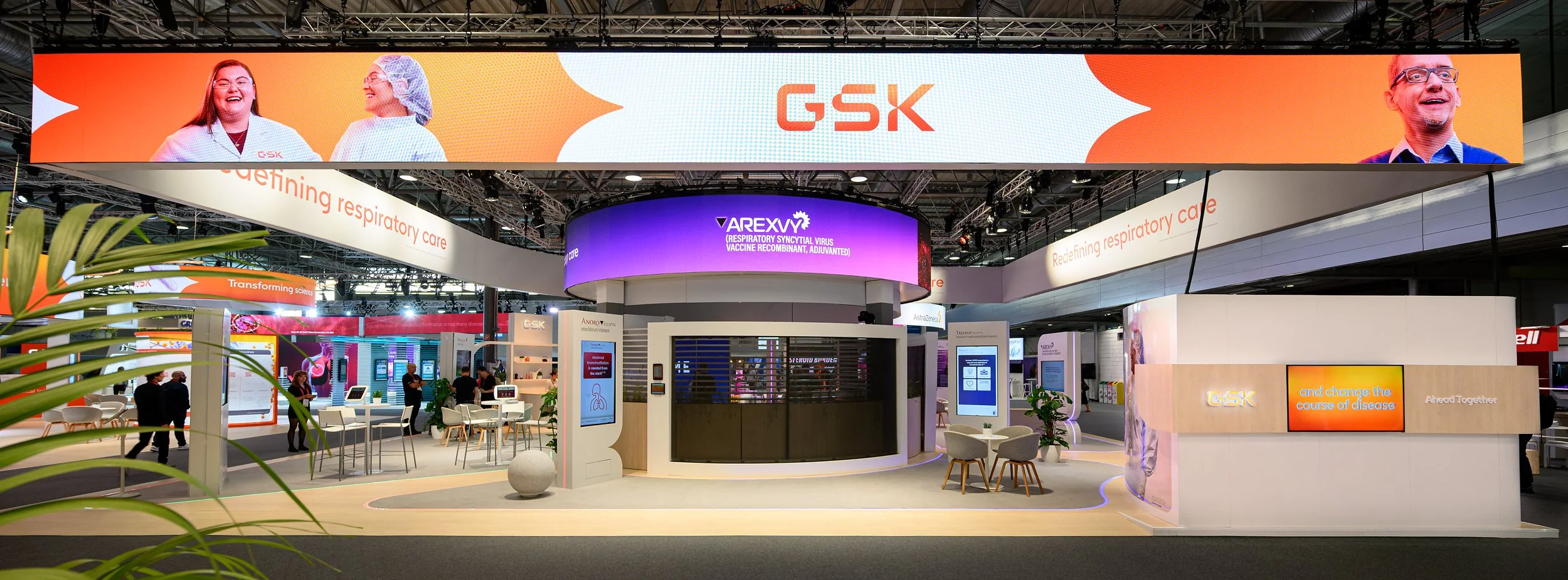 Exhibition booth with GSK branding, large digital screens displaying healthcare-related images and information, and multiple smaller display areas with people engaging at the booth.