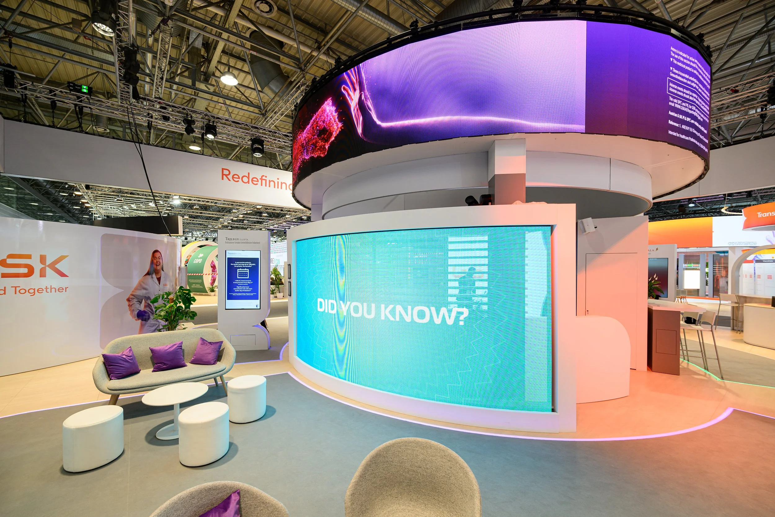 An indoor exhibition area with curved digital screens displaying vibrant graphics and text, modern seating with purple pillows, and booth structures in the background.