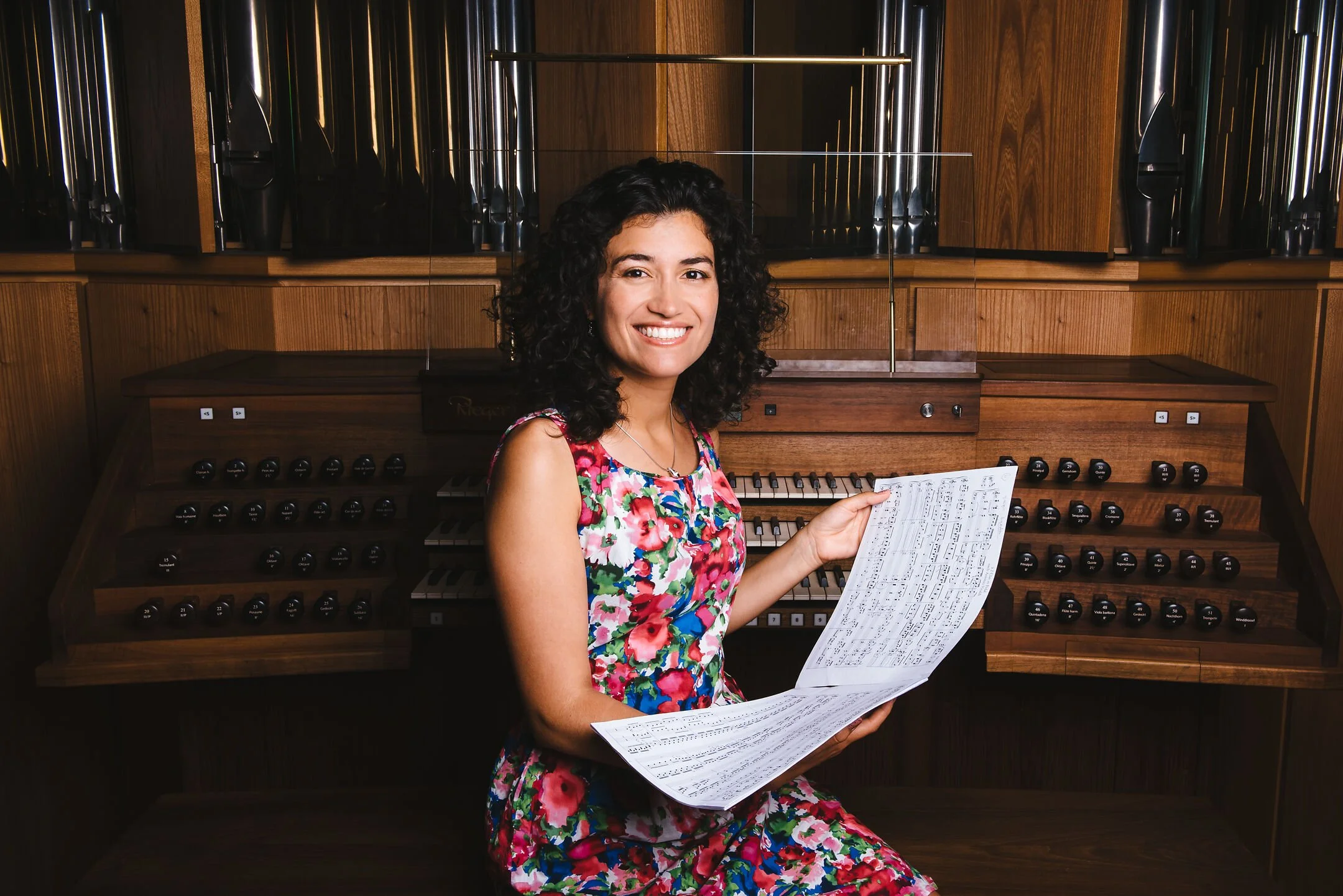 A woman smiling and holding sheet music while sitting at a pipe organ in a wooden room.