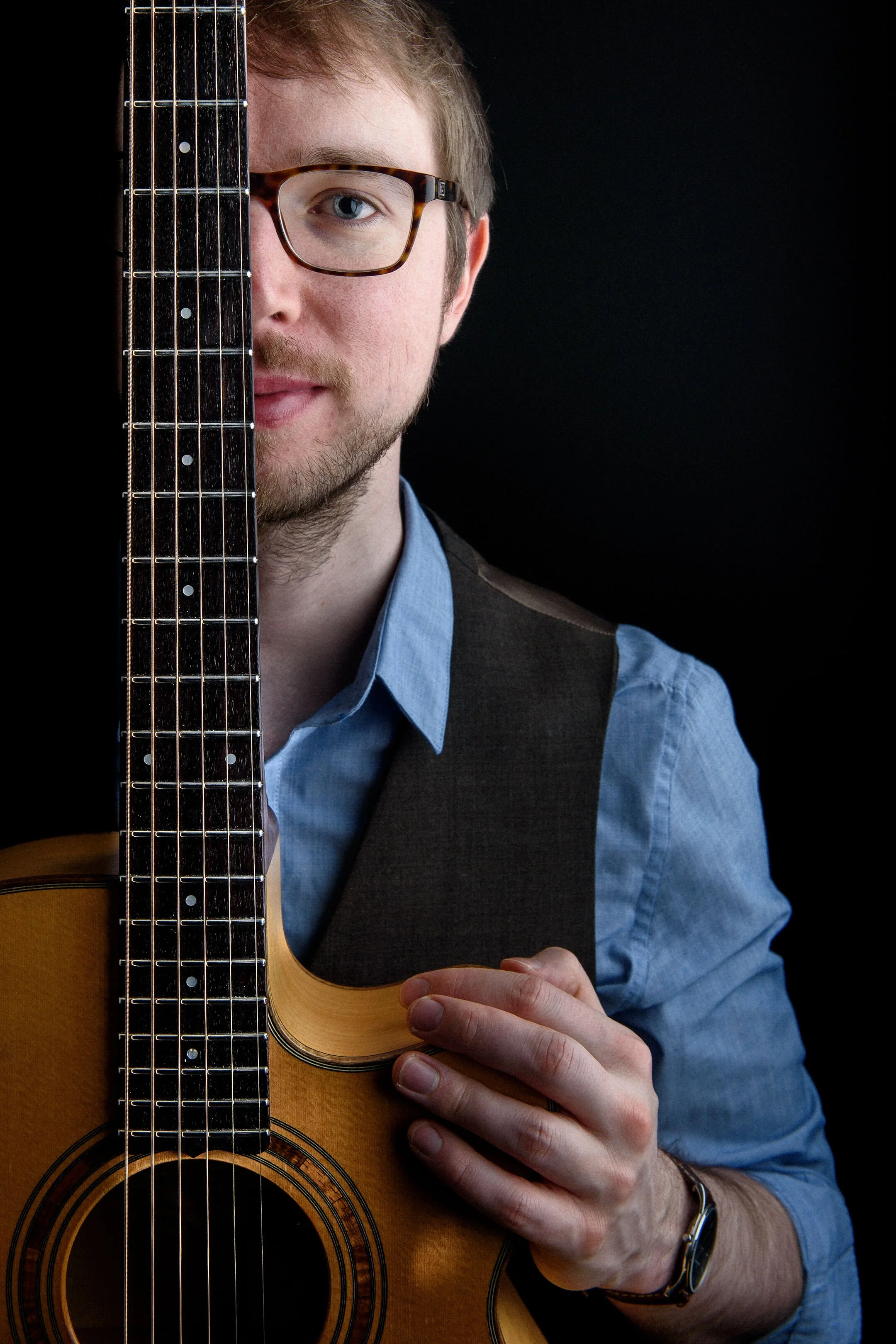 A young man with glasses, a beard, and a blue shirt holding an acoustic guitar, partially hiding his face behind the guitar, against a black background.