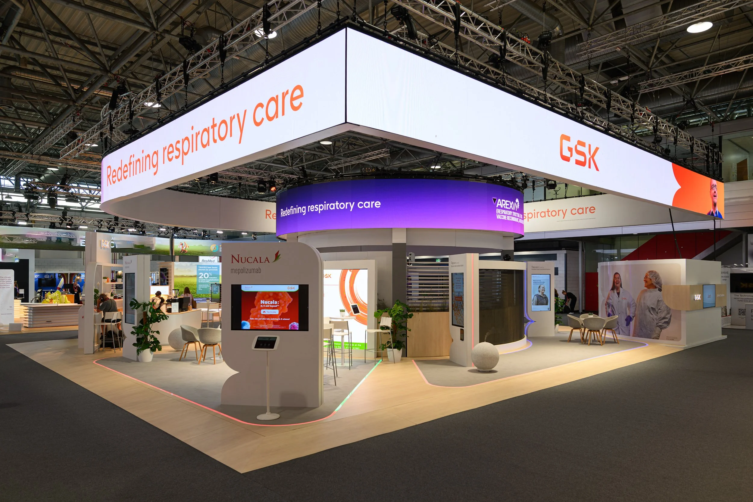 Trade show booth with large LED banner reading 'Redefining respiratory care' and 'GSK,' containing displays, seating areas, and people.
