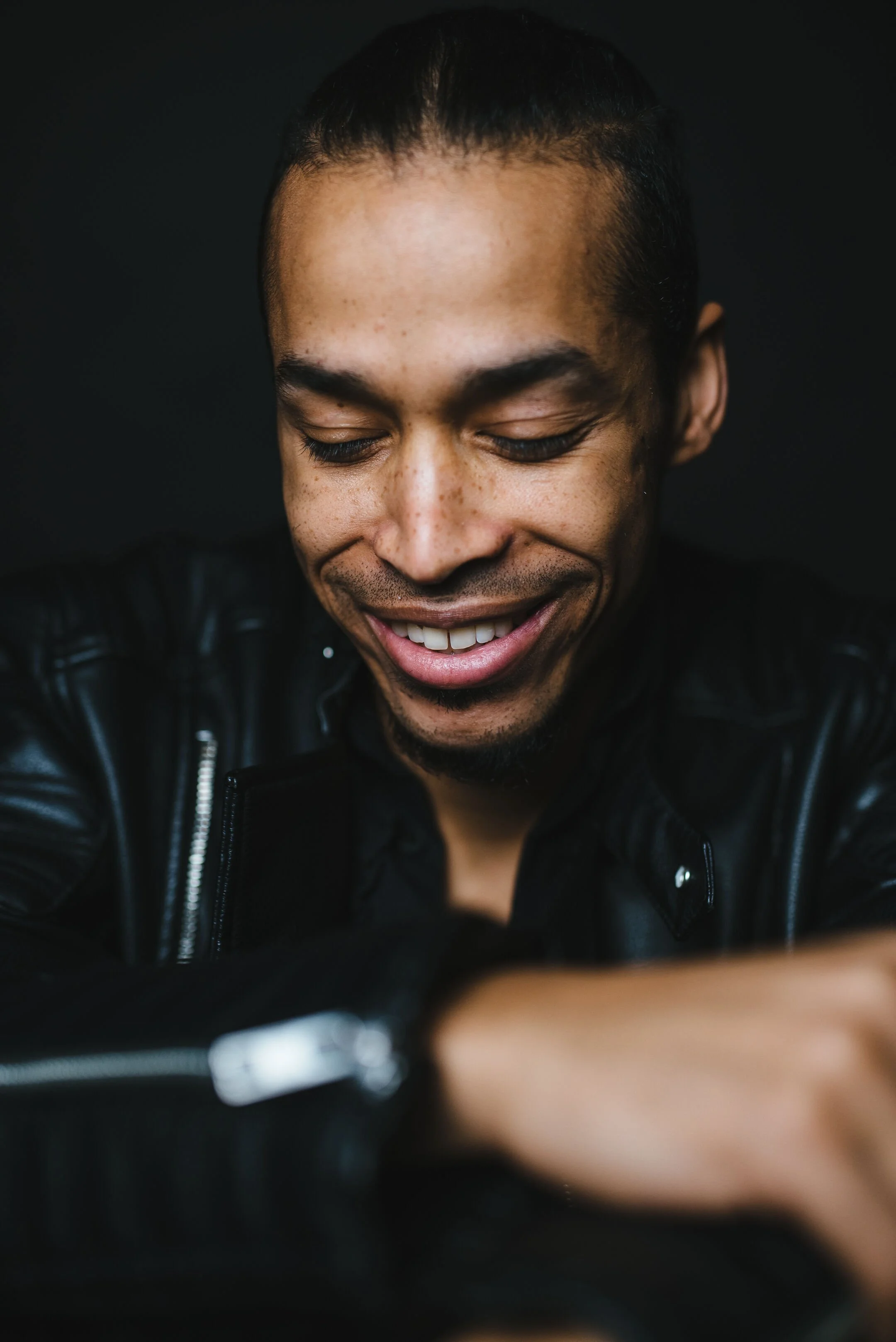 Close-up of a smiling man with short hair and a beard, wearing a black leather jacket, looking down with closed eyes.