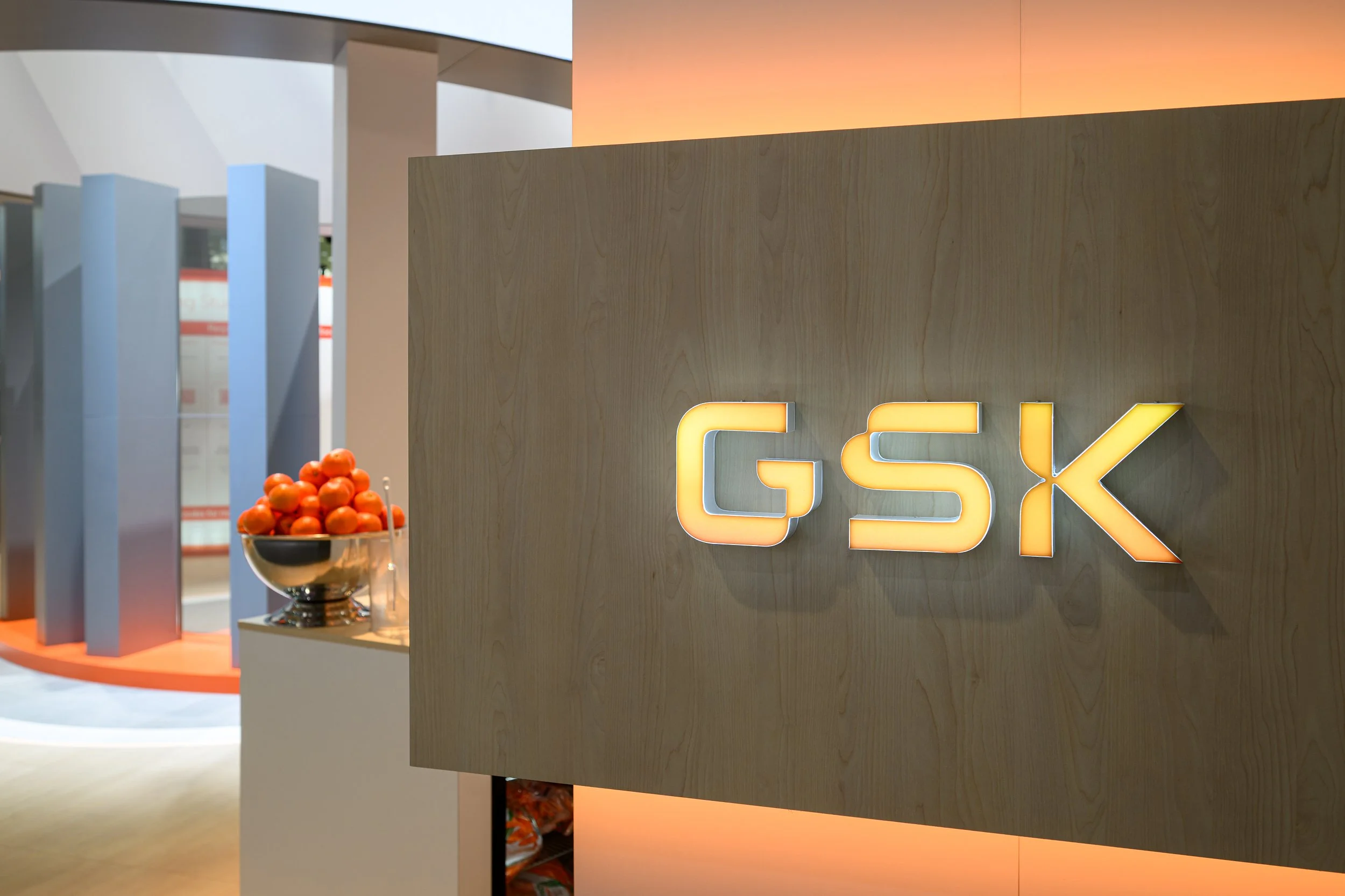 A wooden sign with the text 'GSK' illuminated in yellow, in an indoor setting with a bowl of orange balls on a white surface in the background.