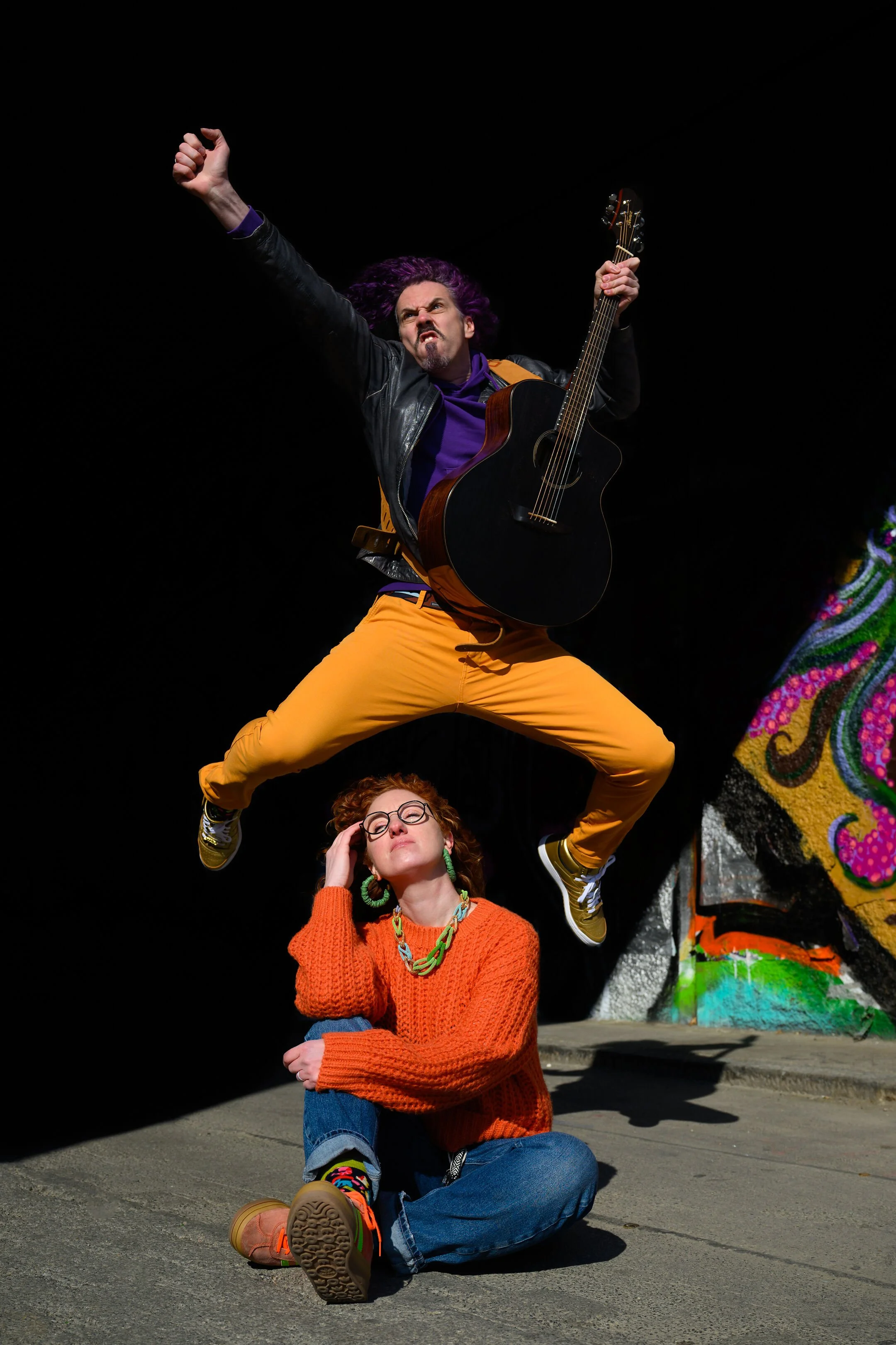 A man is jumping in the air while holding a guitar, with a woman sitting on the ground underneath him. The woman is wearing glasses and a bright orange sweater, and she is sitting with her hand on her head. The background features a black space with 