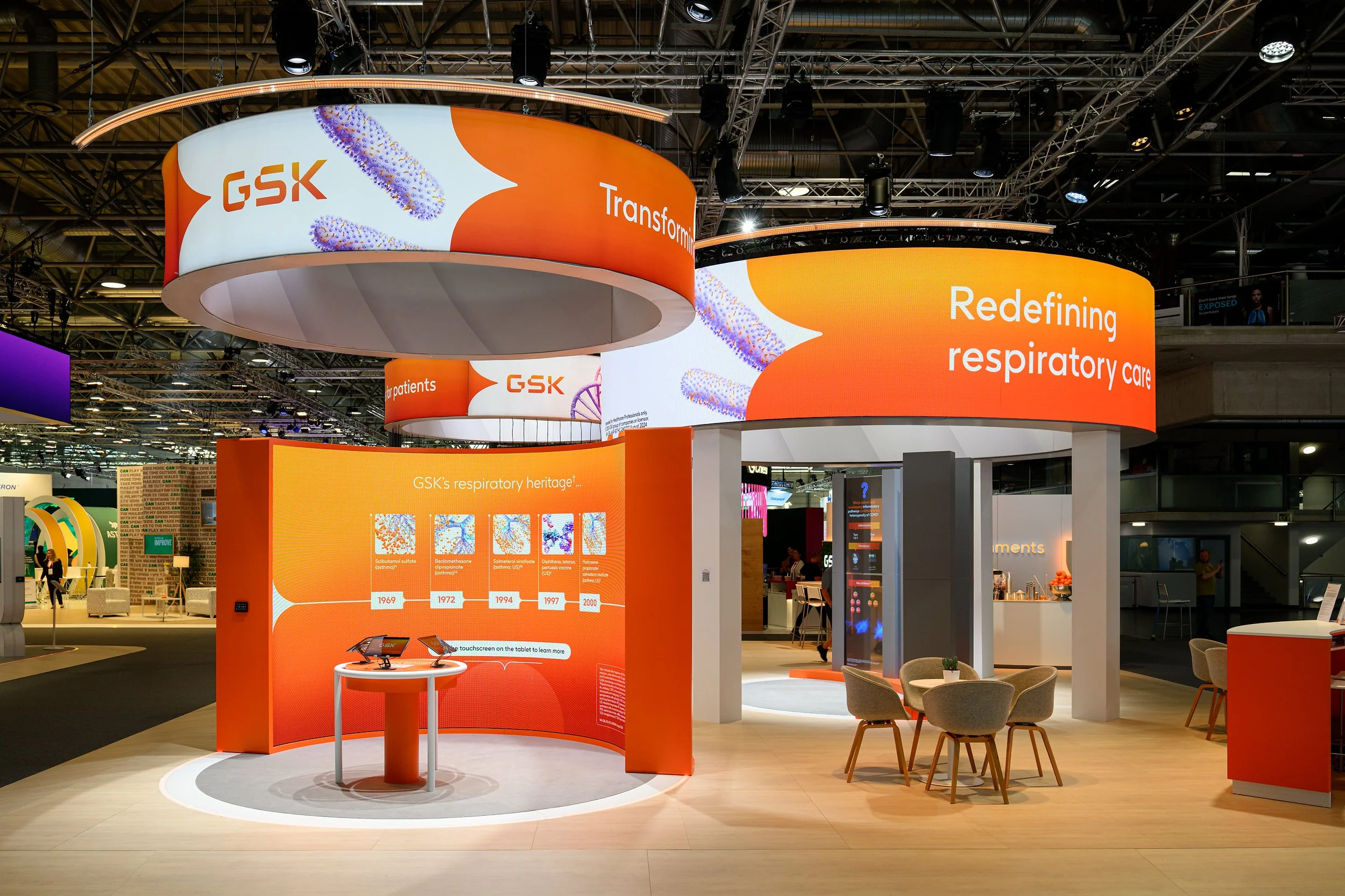 Exhibition booth with large orange and white digital banners displaying the GSK logo and the phrases "Transforming respiratory care" and "Redefining respiratory care." The booth features informational displays, seating area, and interactive tablets, 